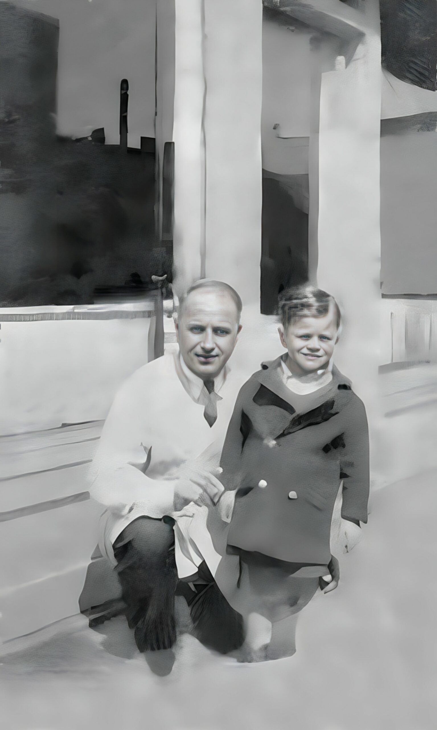 Man and boy posing outdoors in front of a pale modern building; both wearing coats (grayscale, artistic photo).