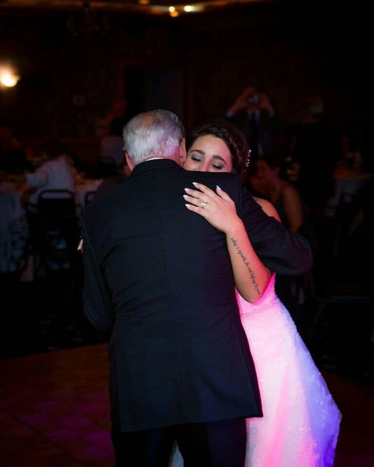 Bride in a white gown hugs an older man in a dark suit on a dimly lit dance floor at a wedding reception.
