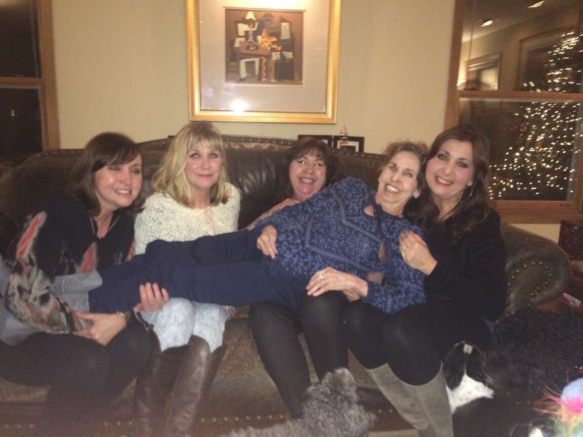 Five women sit on a sofa, smiling as they hold a woman across their laps in a cozy living room with Christmas lights in the background.