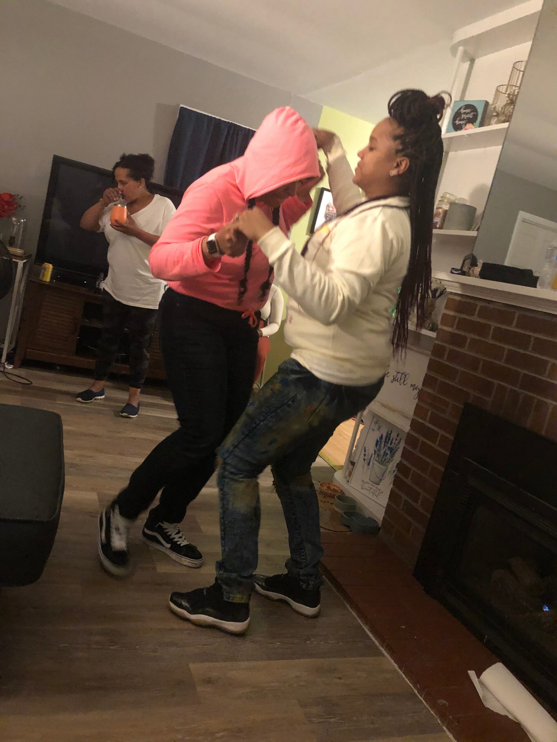 Two people dancing in a cozy living room, one in a pink hoodie with hood up and the other in a white shirt beside a brick fireplace.