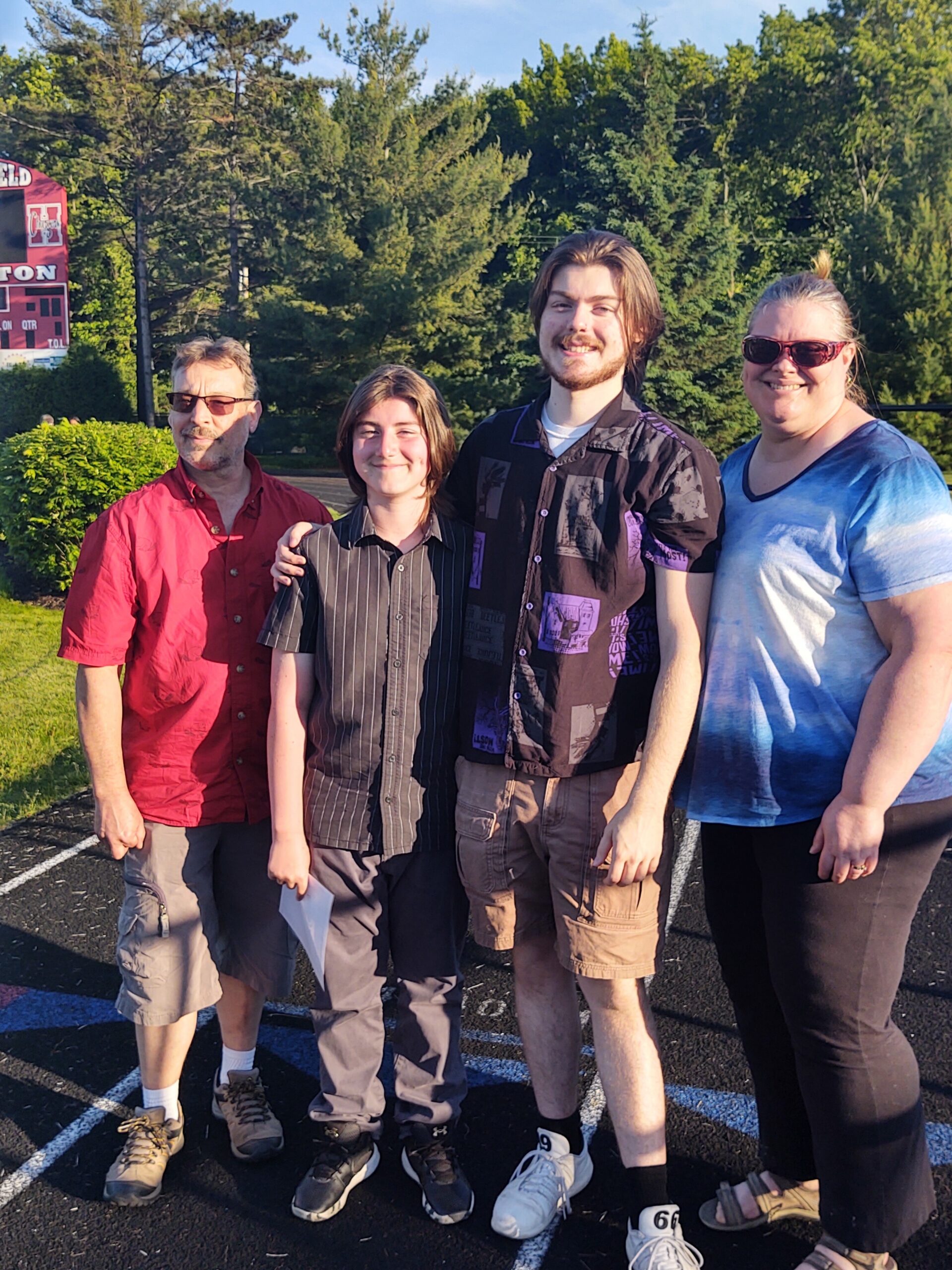 Four people posing together outdoors in a parking lot, smiling with trees in the background (sunny day).