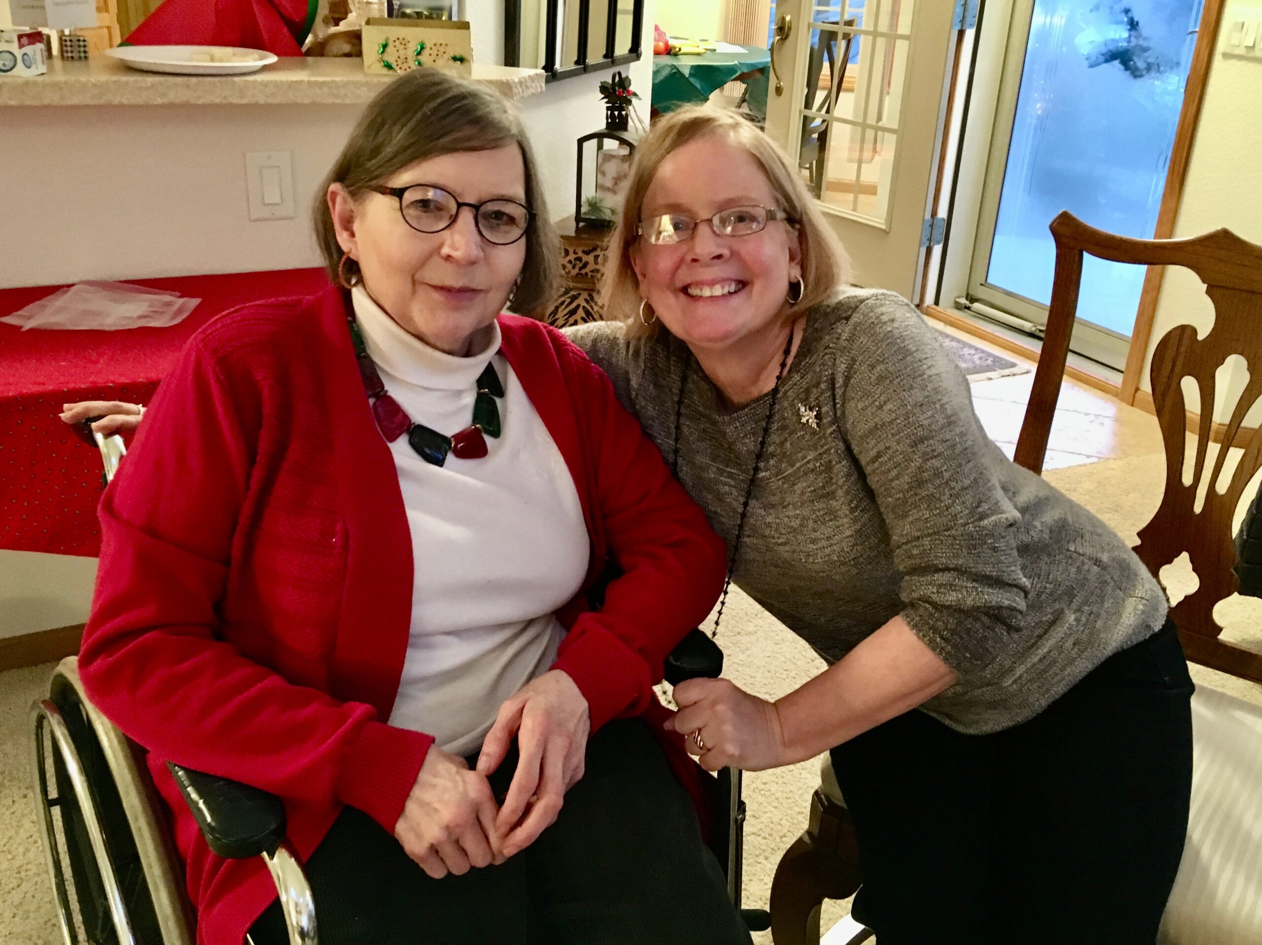 Two smiling women pose together in a home, one in a red cardigan seated in a wheelchair and the other standing beside her in a gray sweater.