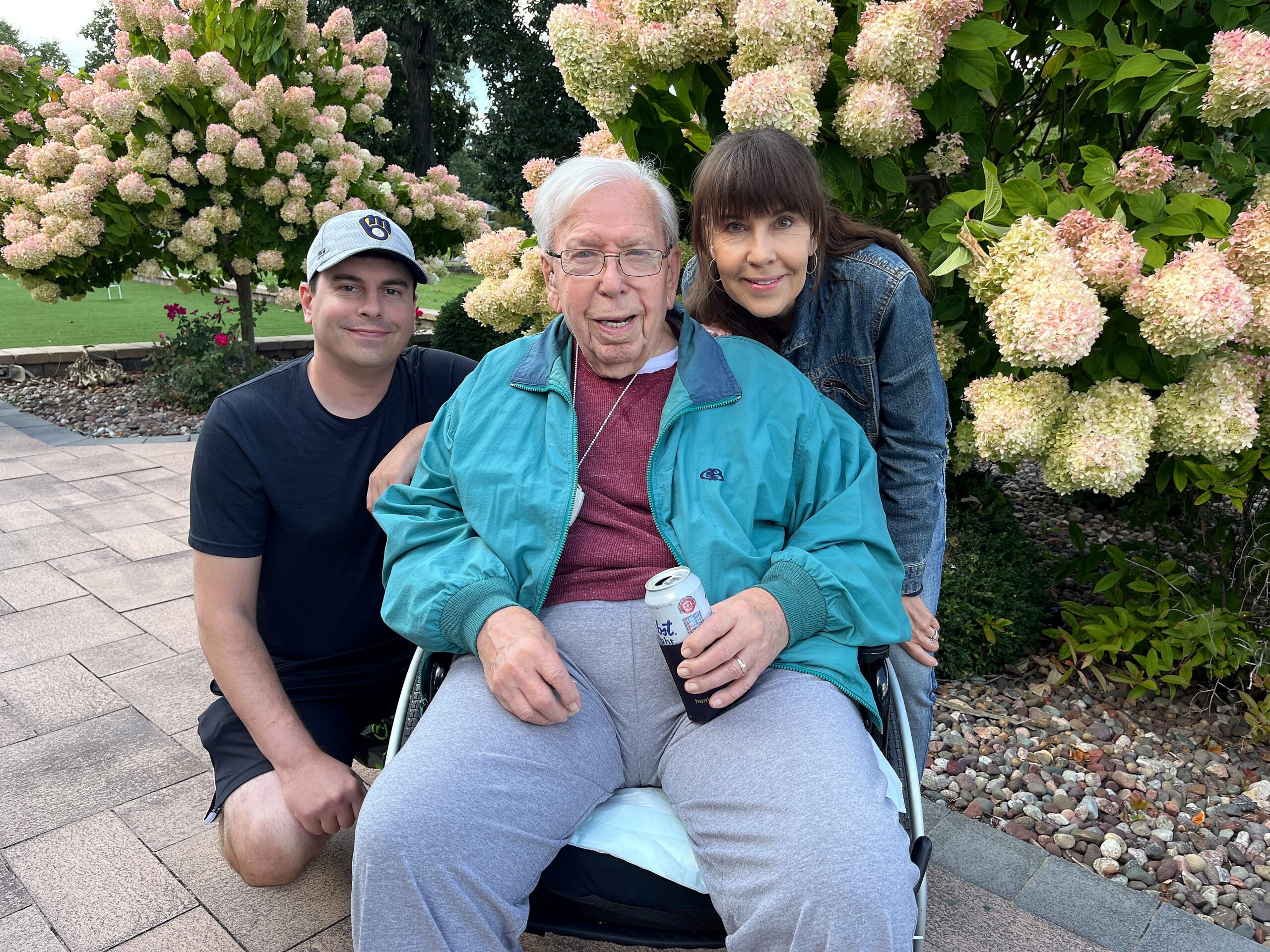 Three people posing outdoors in front of pink and white hydrangea bushes: an elderly man in a wheelchair center, with a younger man on his left and a woman leaning over from the right.