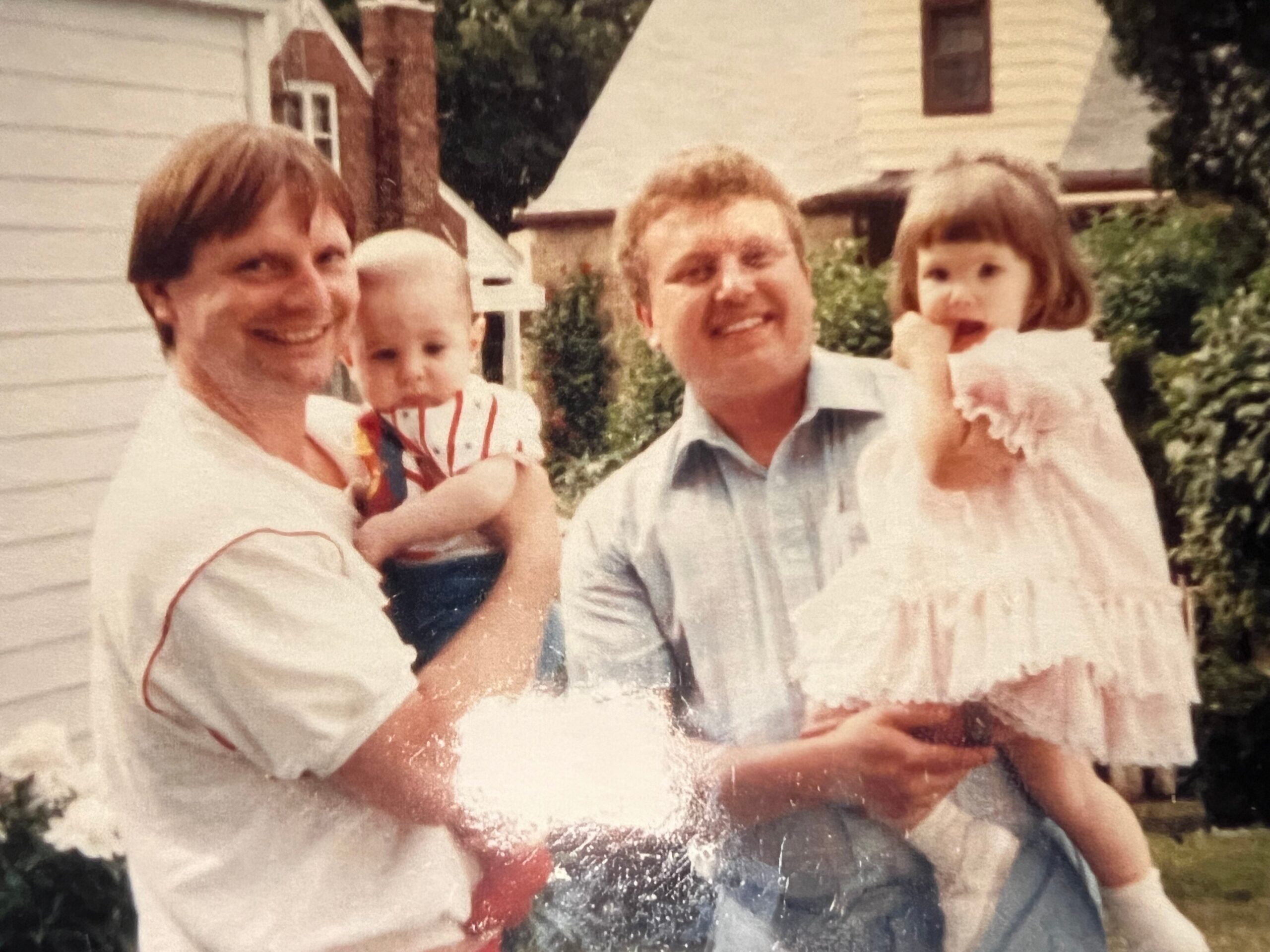 A family photo outdoors: two men and two young children posing together, with a house and greenery in the background.