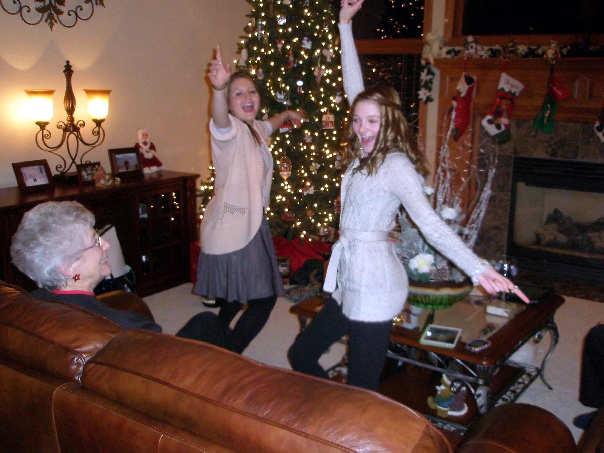 Two young women dancing and smiling by a warmly lit Christmas tree in a cozy living room, while an elderly woman sits on a leather couch nearby.