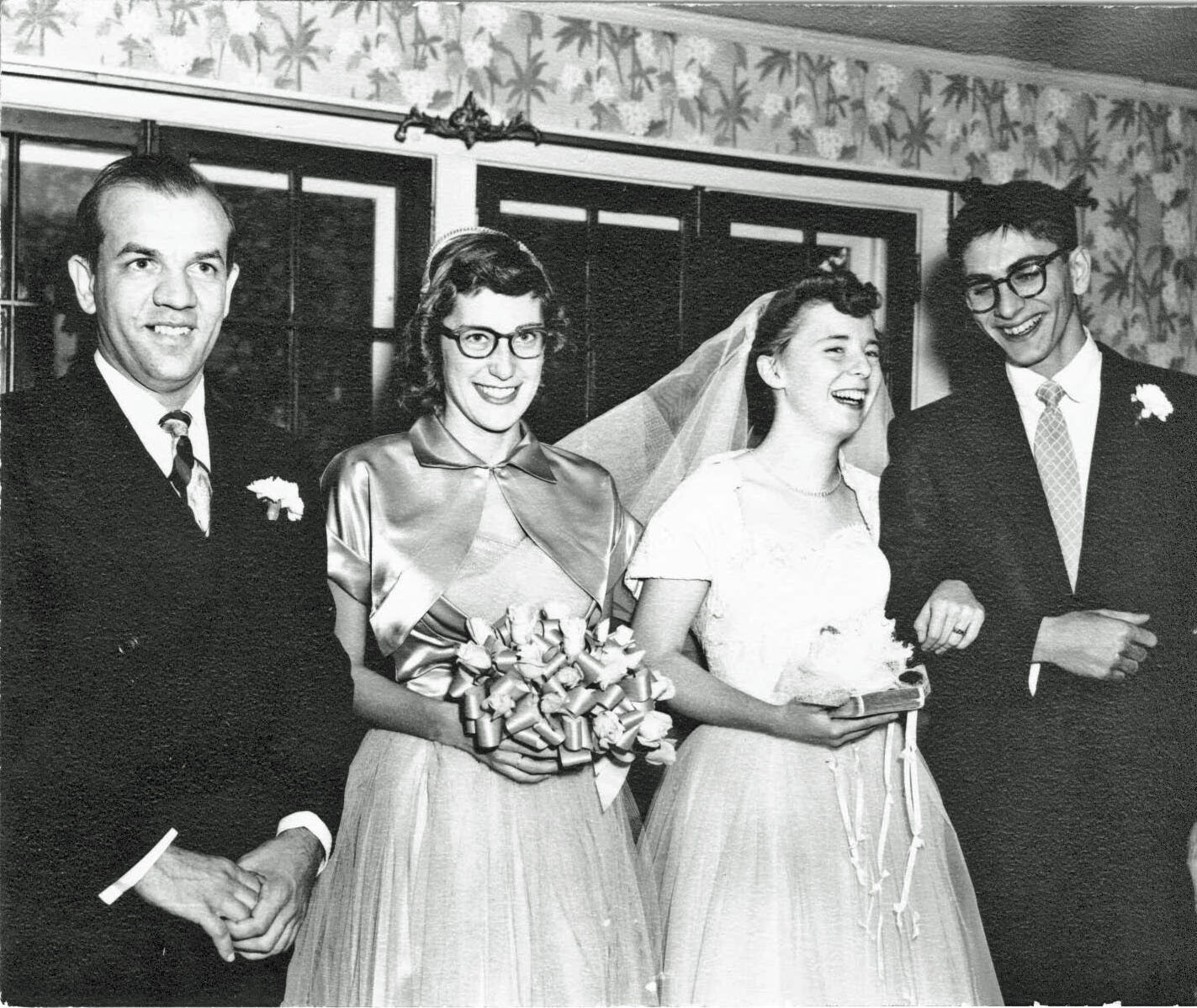 Four people at a wedding: two brides in white dresses (one with veil, one with a satin cape) flanked by two men in dark suits, all smiling indoors.