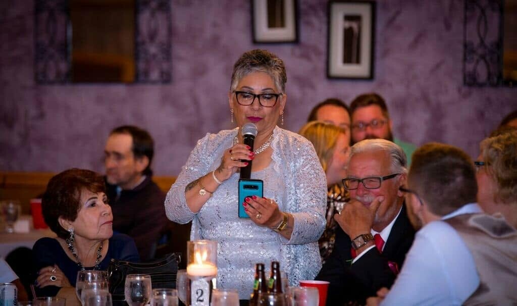 Woman in silver blouse speaks into a microphone at a dinner table, holding a blue phone as guests listen nearby.