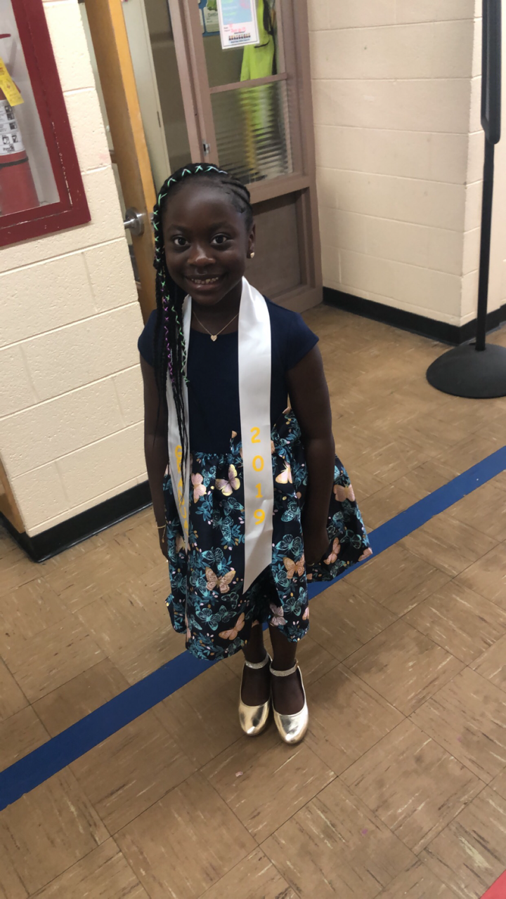 Smiling young girl in a butterfly-print dress with a white 2019 sash, standing in a school hallway in gold shoes.
