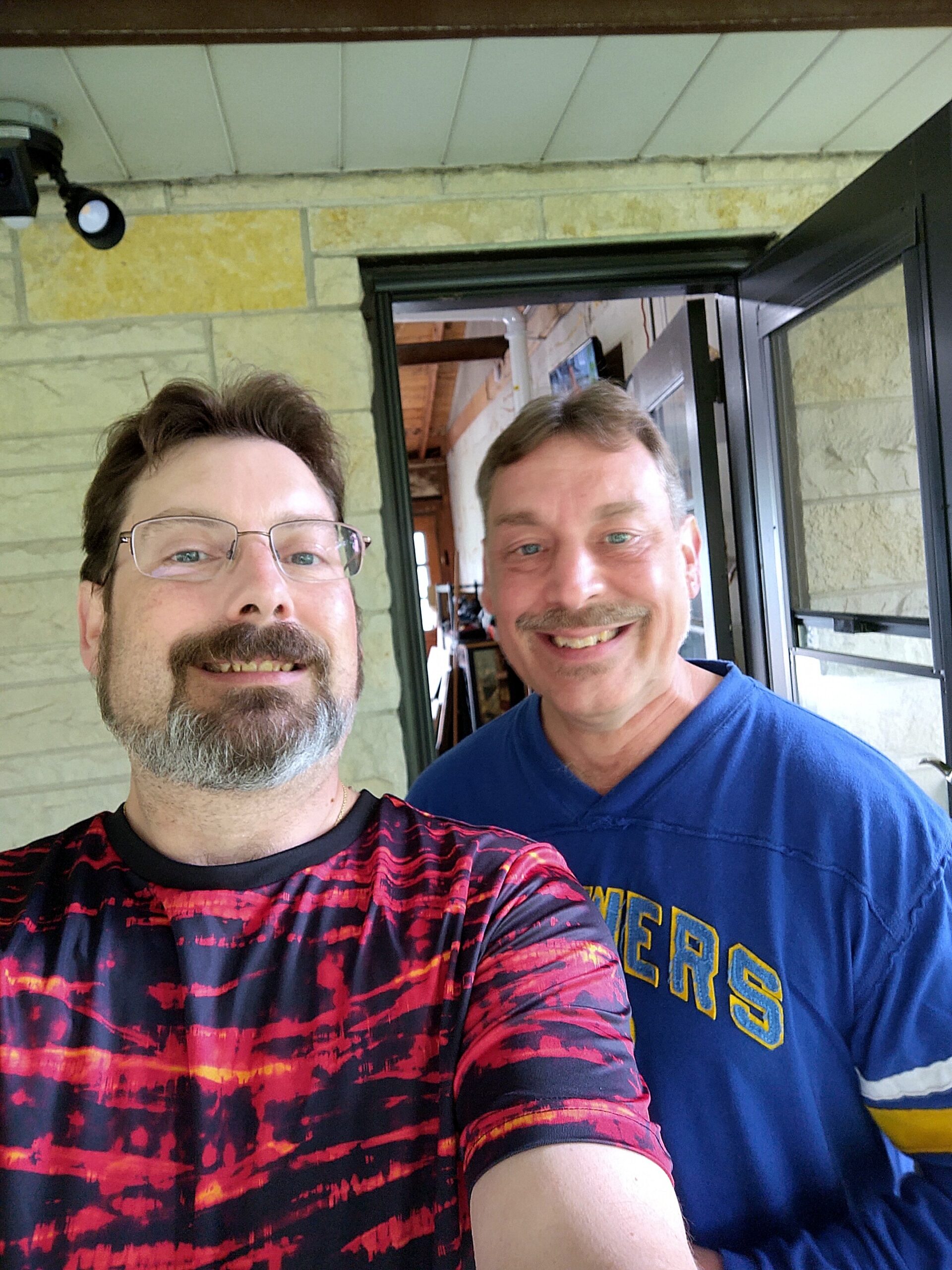 Two men pose for a selfie on a porch: one in a red-black patterned shirt in the foreground, the other in a blue sweatshirt behind him, both smiling.