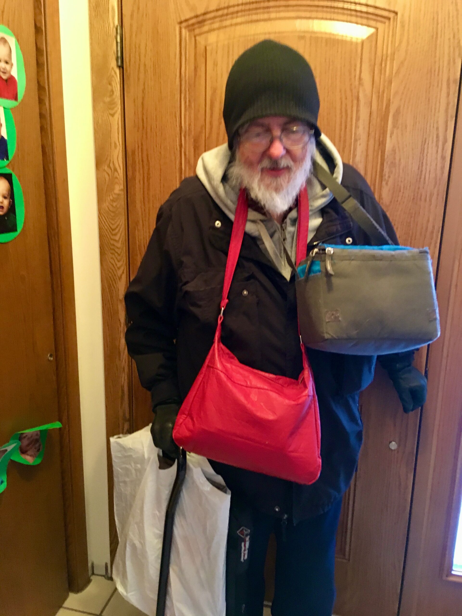 Older man with a white beard wearing a black beanie and glasses, holding a gray shoulder bag and wearing a red apron in a doorway.