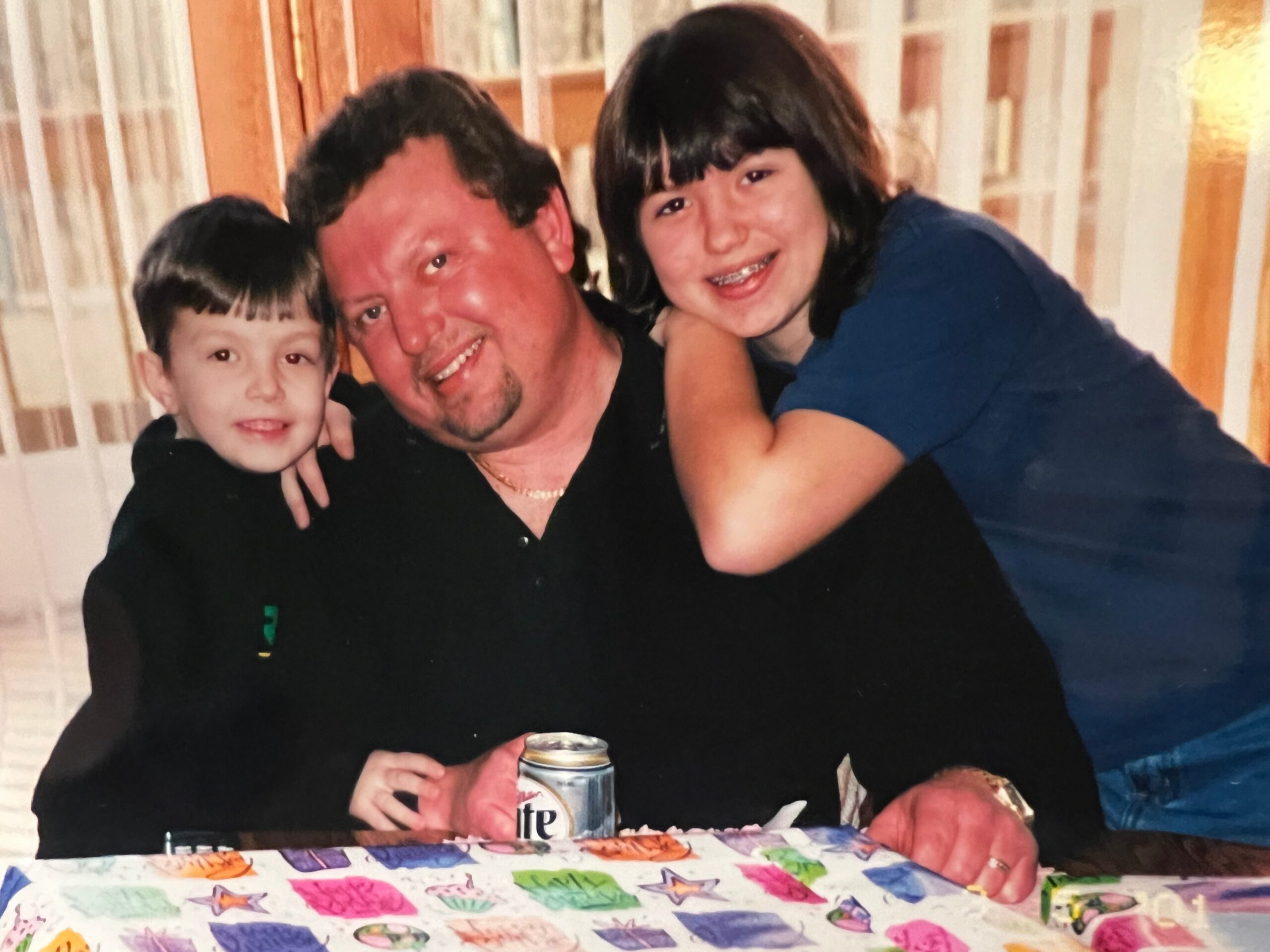 Family photo: a man with two kids leaning in and smiling, at a table covered with a bright, patterned tablecloth; a beer can is visible in the foreground.