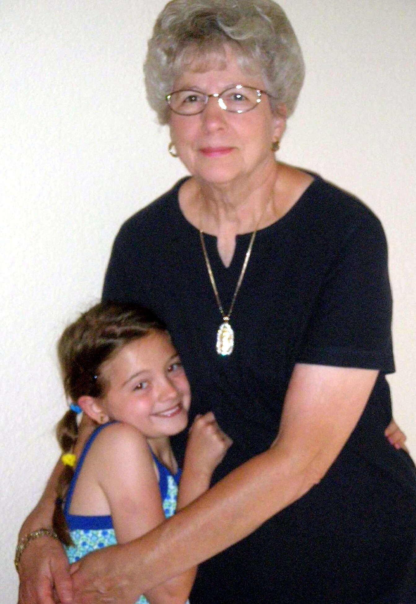 Older woman with short gray hair and glasses wearing a black top and a pendant, embracing a smiling young girl in a blue dress.