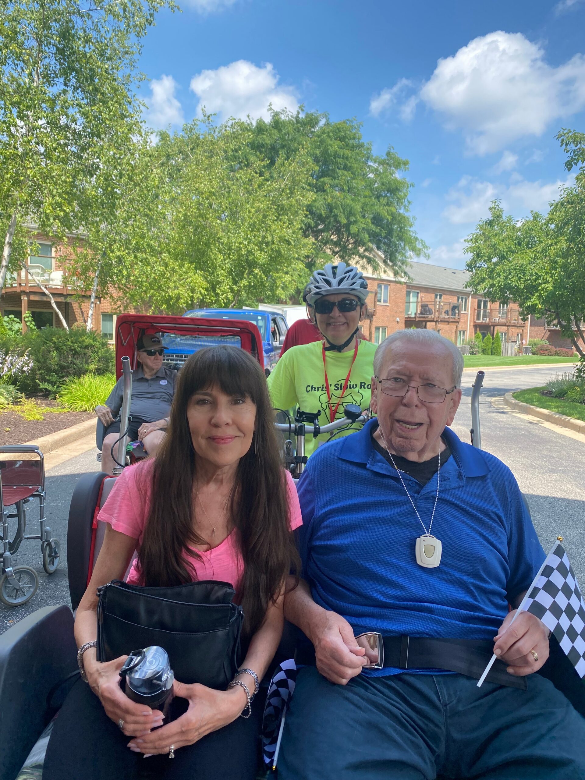 Smiling woman in pink top sits in a wheelchair-like chair next to a man in a blue polo shirt on a sunny residential street with trees and brick buildings behind them, holding a checkered flag nearby.