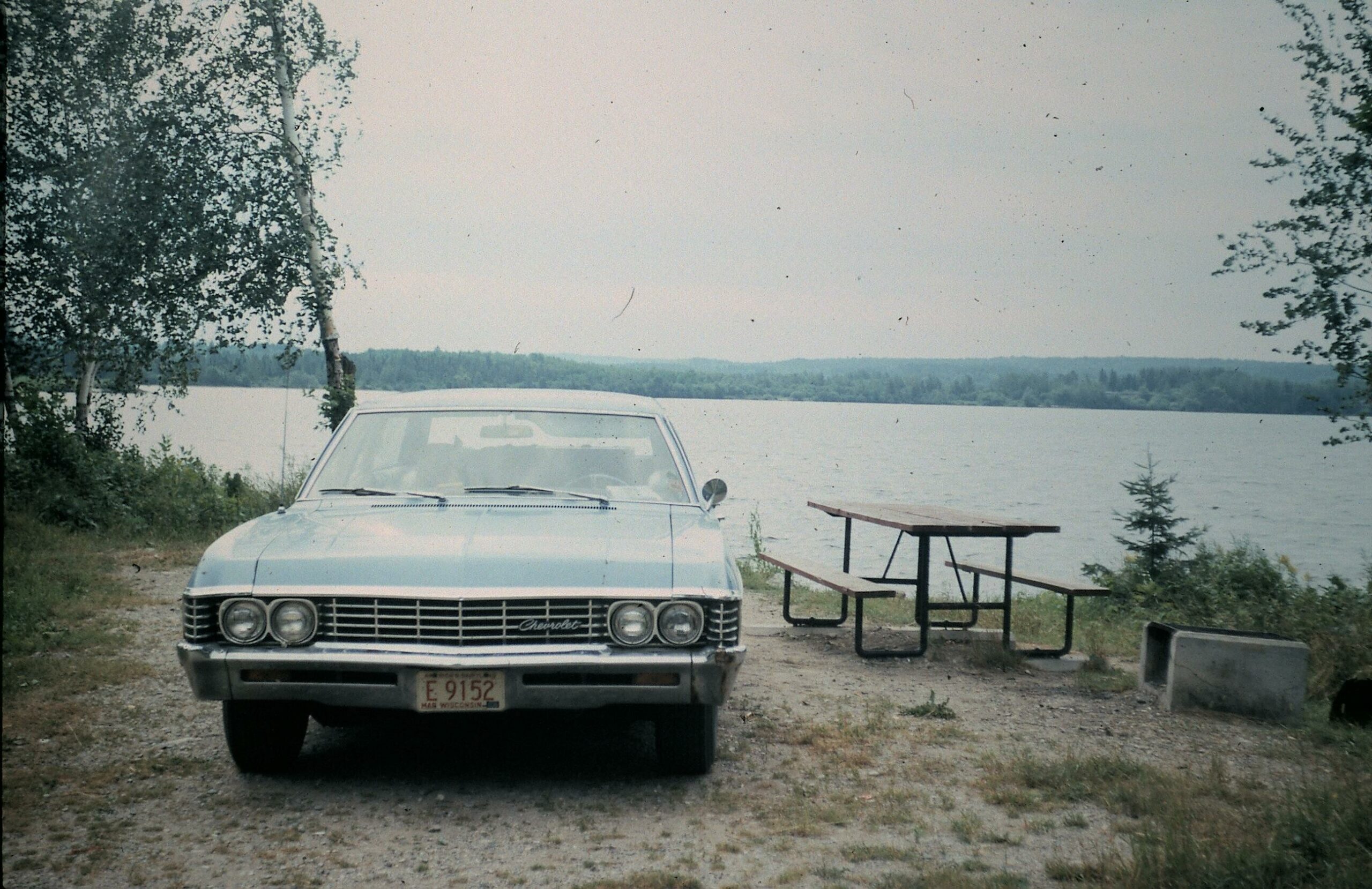 Front view of a light blue vintage Chevrolet parked on a dirt area by a lake, with a wooden picnic table to the right and trees in the background.