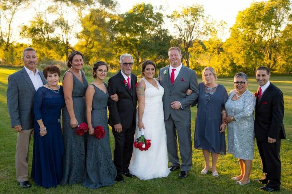 Bride in a white dress and groom in a gray suit pose with their wedding party on a sunny lawn.