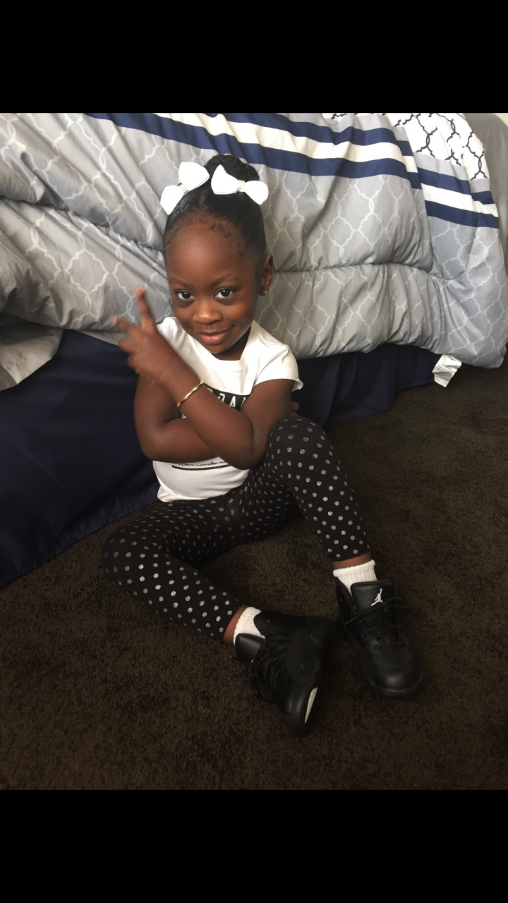 Young girl sitting on the carpet next to a bed, making a peace sign with crossed arms, smiling at the camera, wearing a white shirt, polka-dot pants, white socks, and black sneakers with a white bow in her hair.