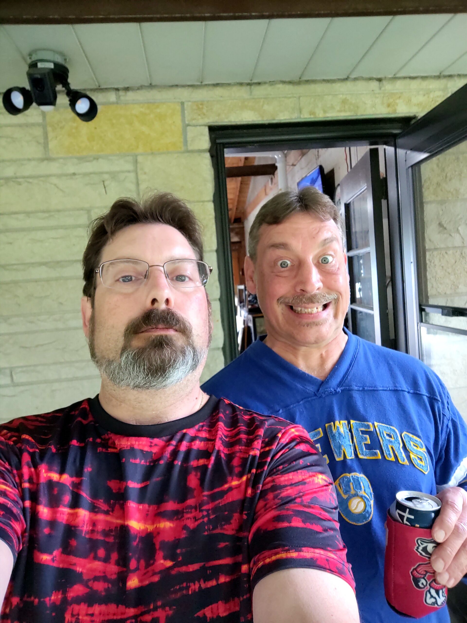 Two men take a casual selfie on a porch or entryway; one in a red and black tie-dye shirt is in the foreground, the other in a blue shirt holding a beer can with a red-cooler sleeve visible; both smile at the camera.