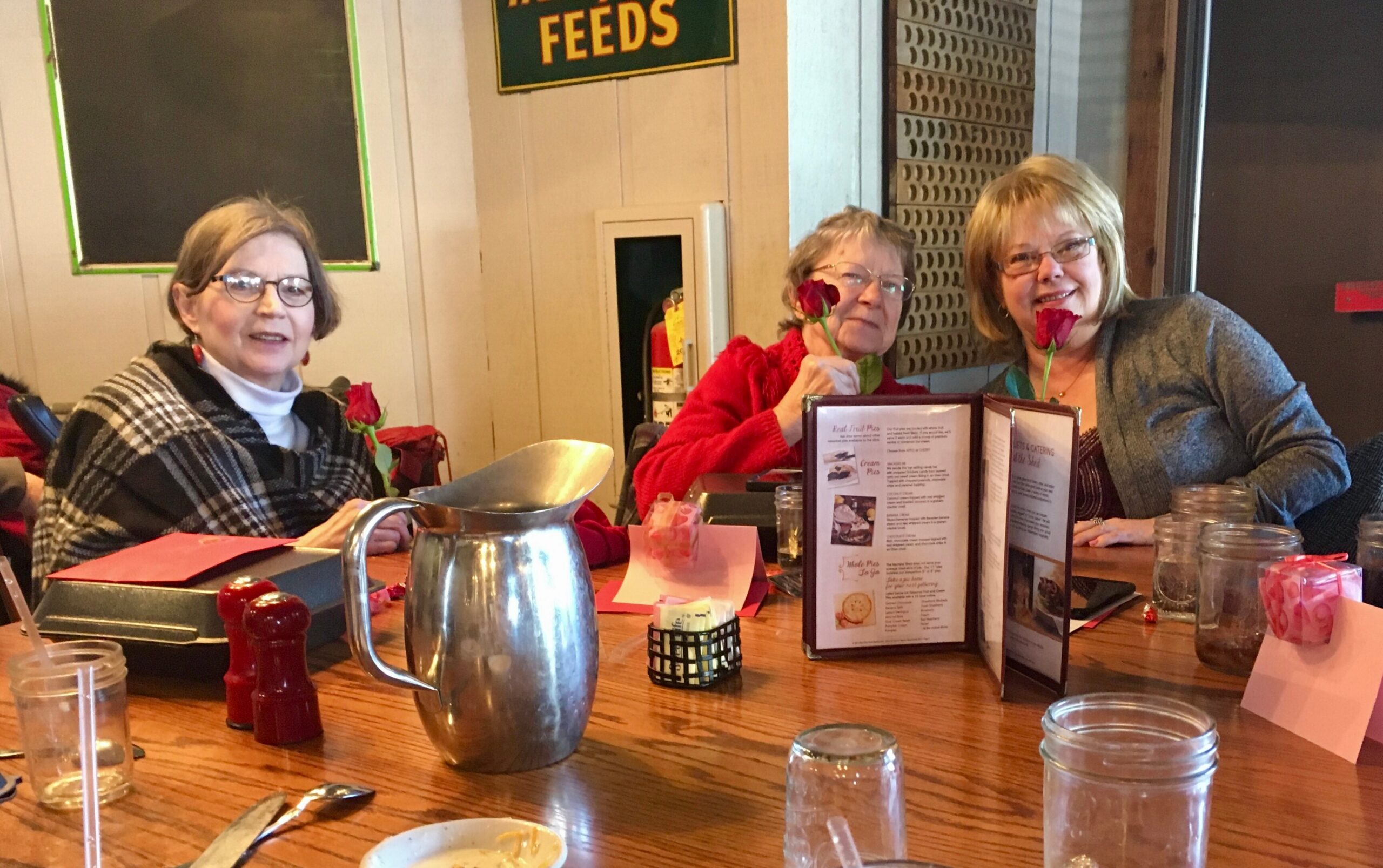 Three women sit at a restaurant table with menus, pink napkins, and roses, posing for a photo.