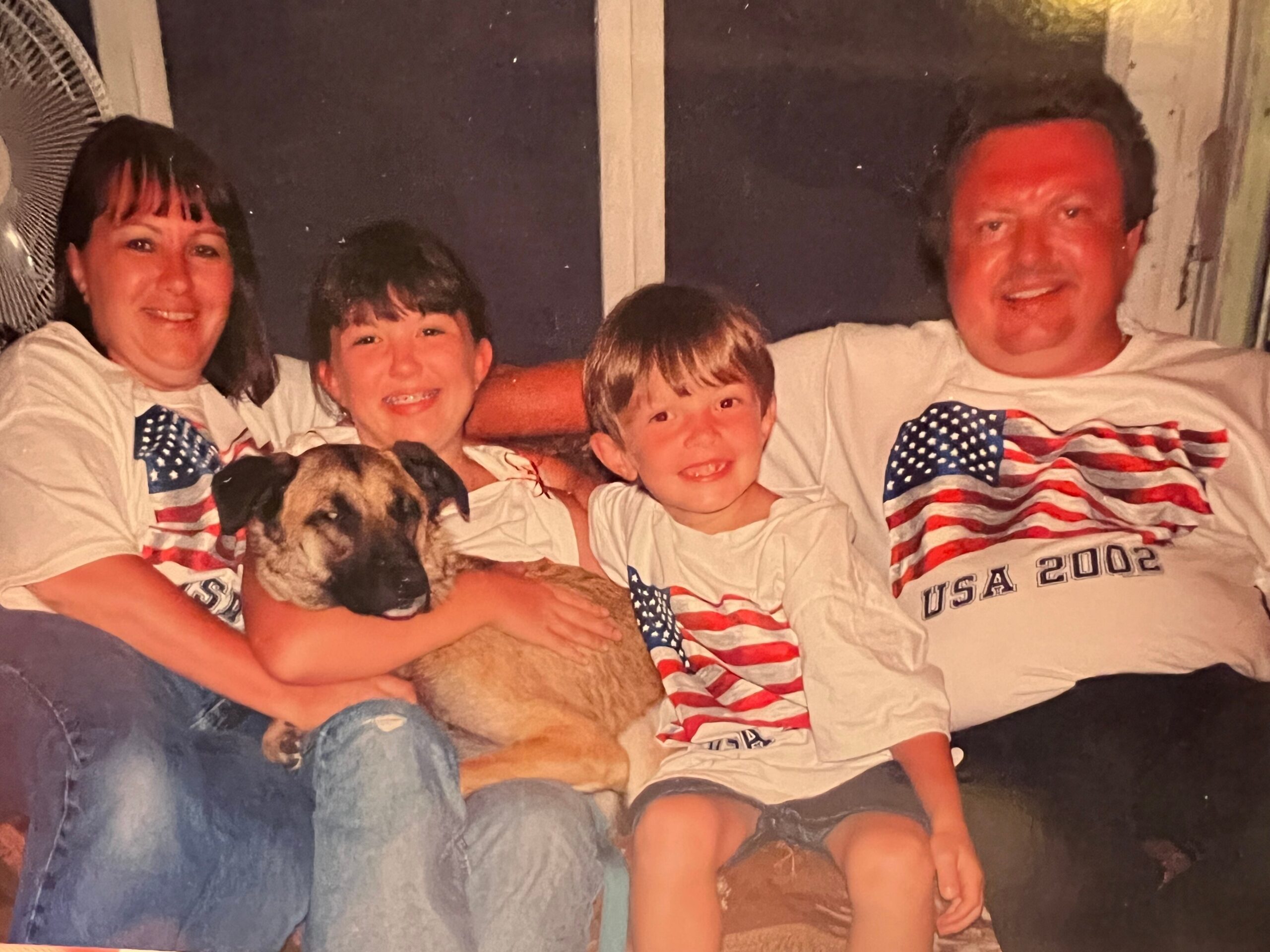 Family group on a sofa wearing matching USA 2002 shirts, with a dog cuddled in a child’s arms, smiling at the camera.
