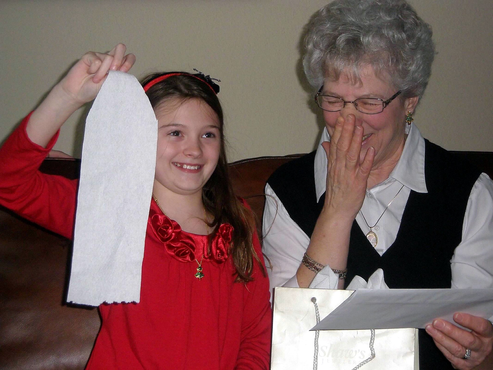 Smiling young girl in a red dress holds up a white napkin next to an elderly woman with gray hair and glasses, who covers her mouth in laughter.