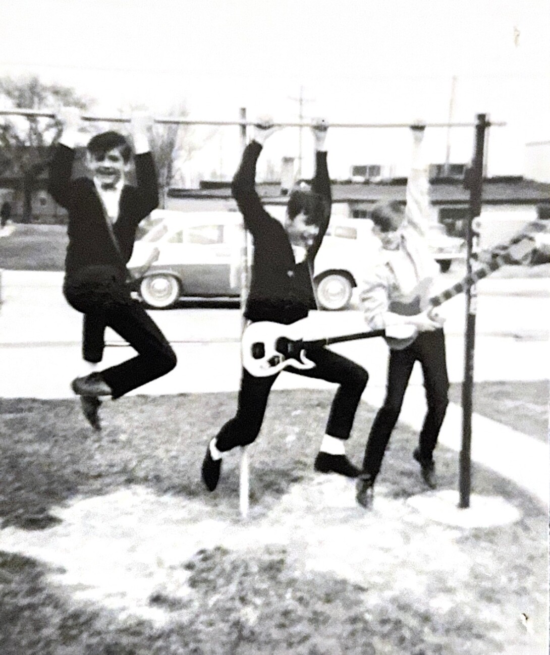 Three young men jump mid-air with arms raised on a grassy park, one holding a white guitar prop.
