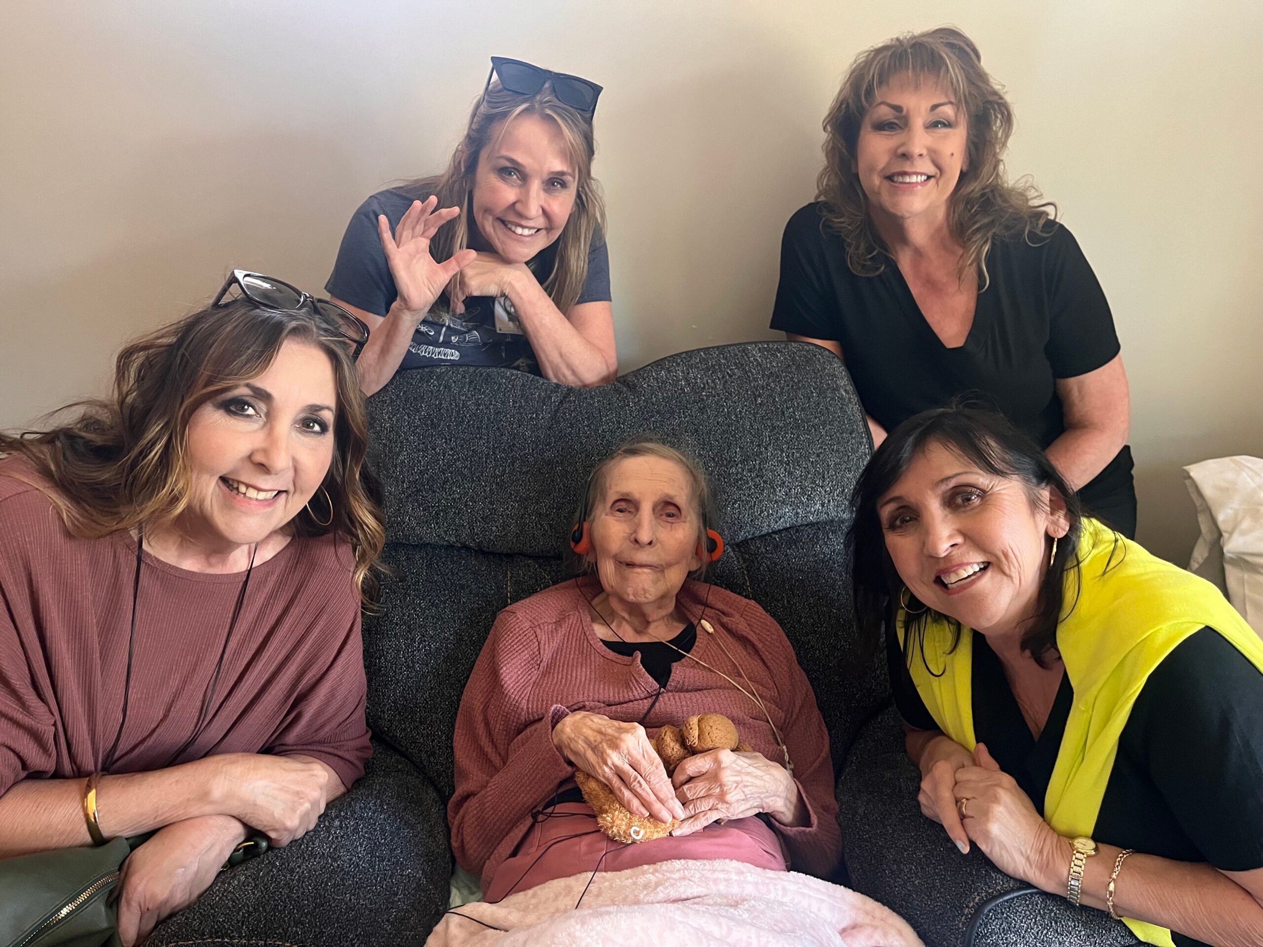 Group of smiling women of different ages sitting on a couch, with an elderly woman in the center holding cookies and snacks nearby.