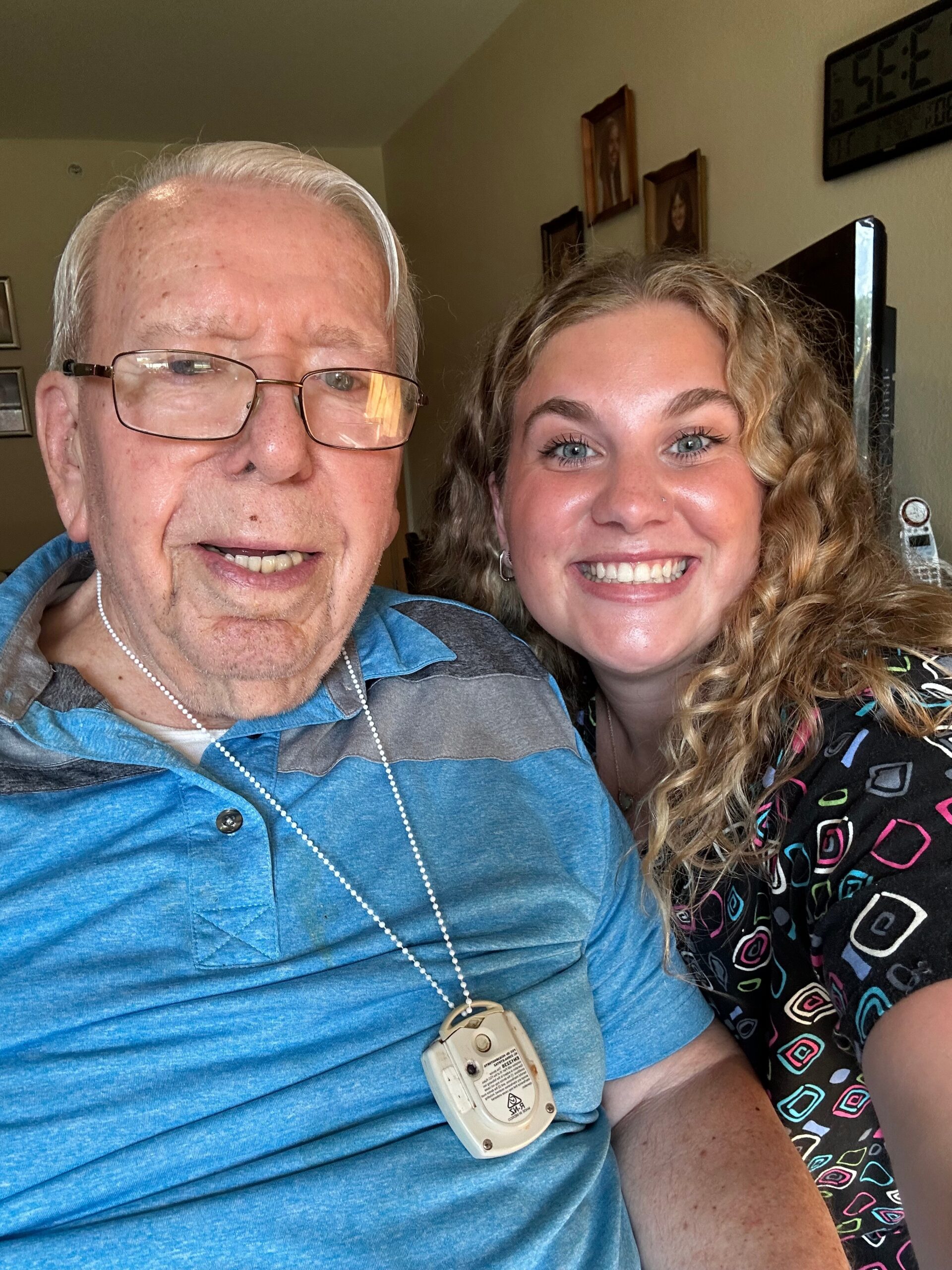Selfie of an older man wearing glasses and a blue striped shirt with a medical alert pendant, smiling next to a young woman with long curly hair indoors.