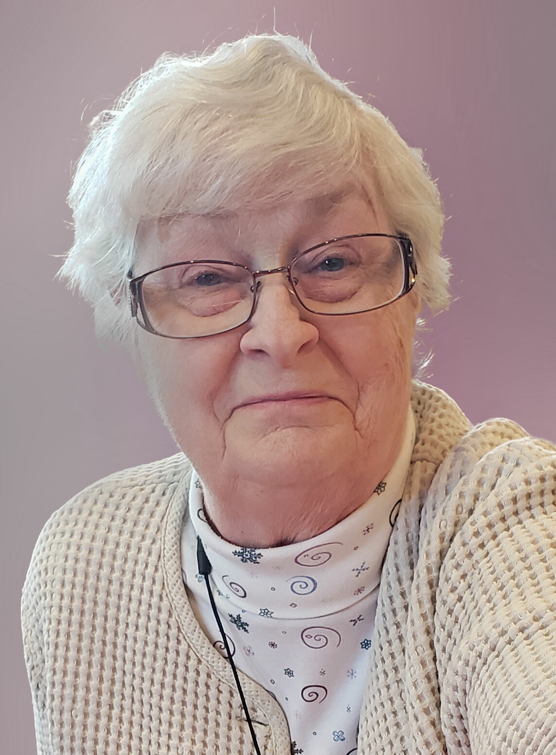 Close-up portrait of an elderly woman with short white hair and glasses, wearing a cream knit cardigan and patterned scarf against a lavender background.