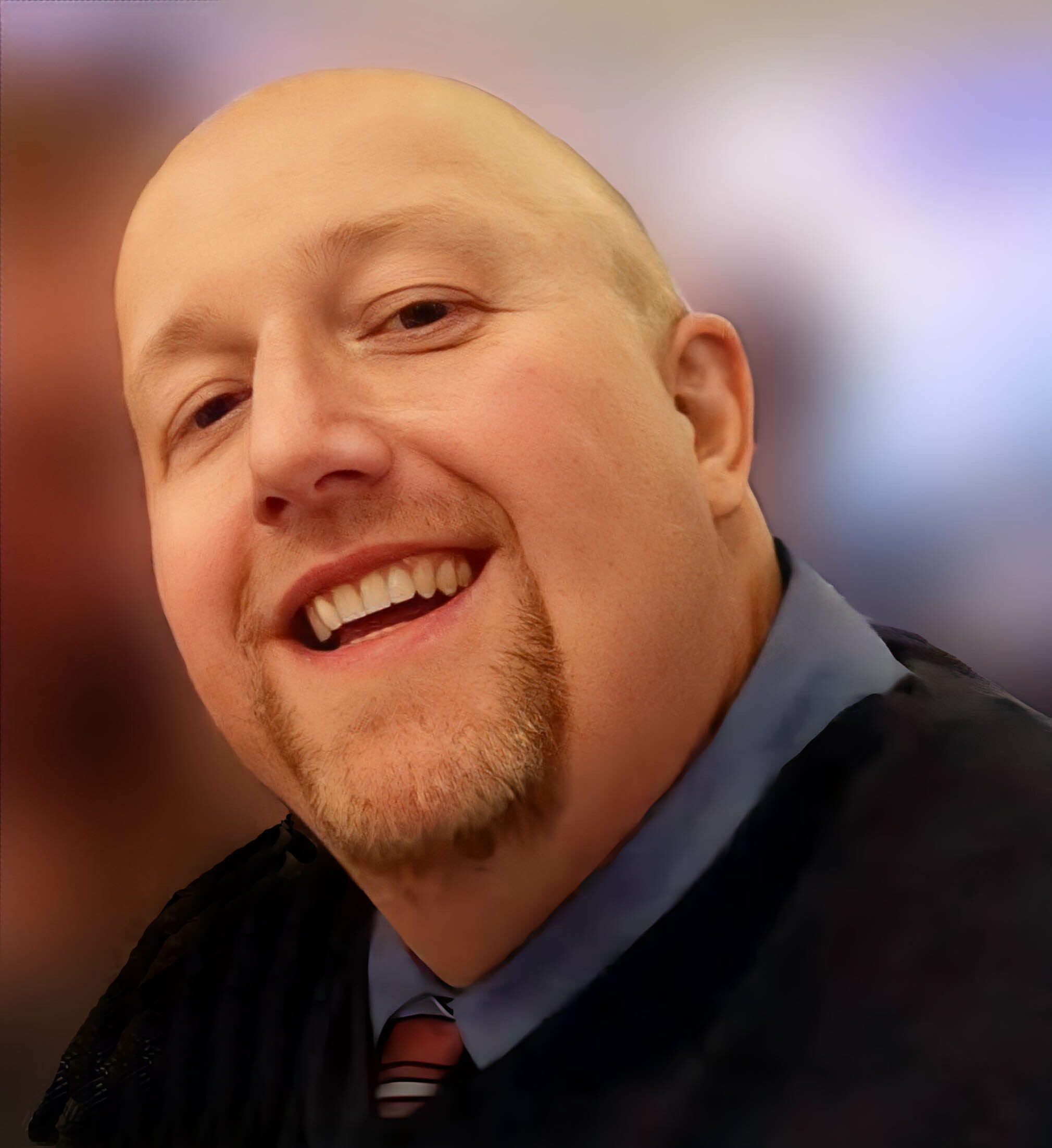 Close-up of a smiling man with a shaved head wearing a navy suit and striped tie, against a blurred background.