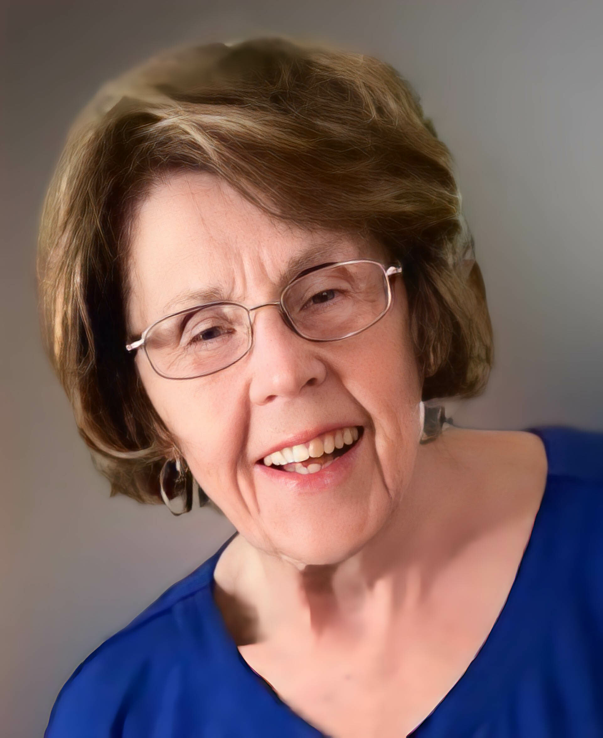 Smiling elderly woman with short brown hair and glasses wearing a blue top.