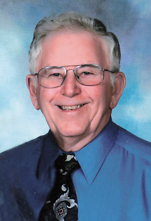 Smiling elderly man with glasses wearing a blue shirt and patterned tie, studio portrait.