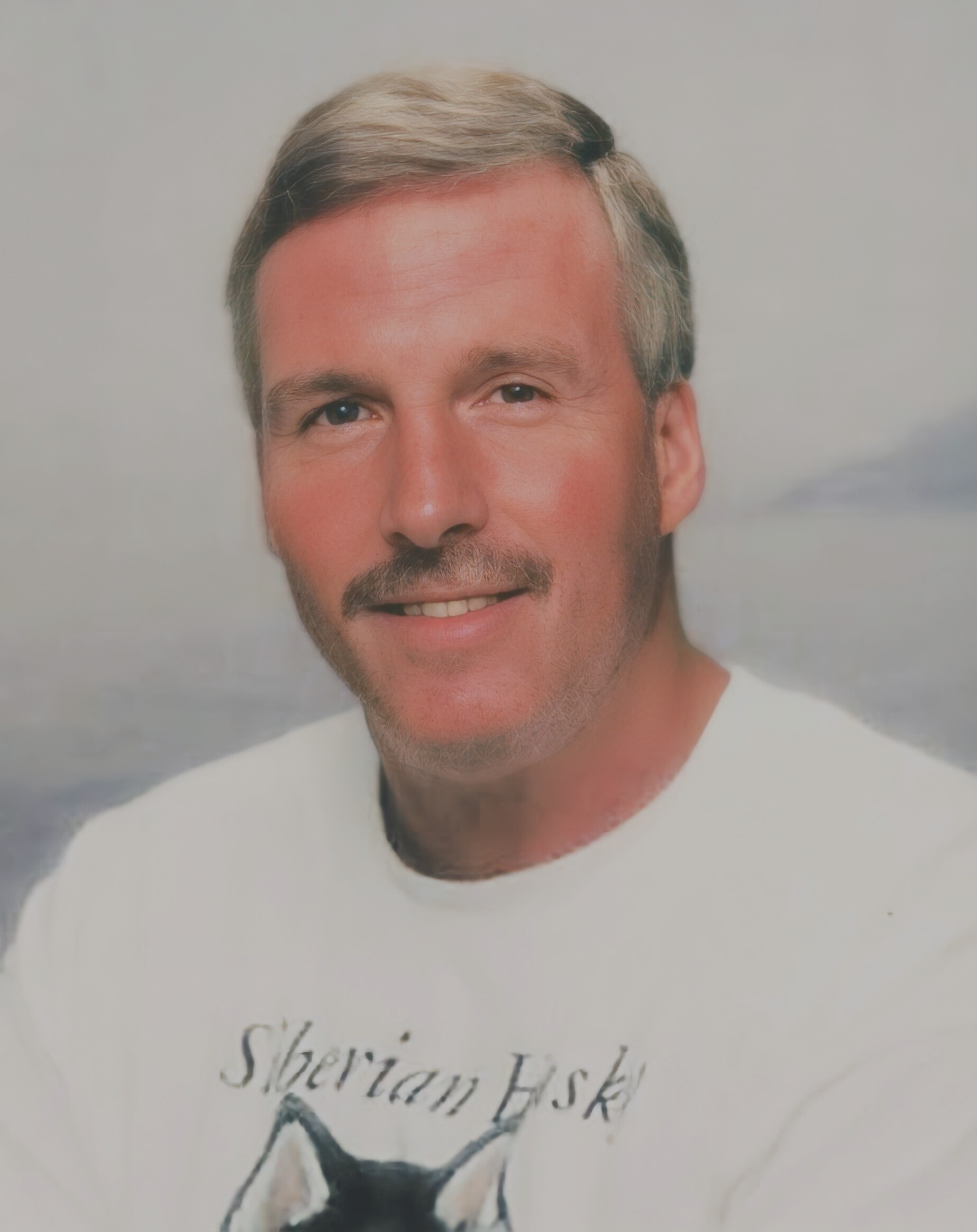 Close-up portrait of a smiling man with blond hair and light stubble, wearing a white shirt against a pale background.