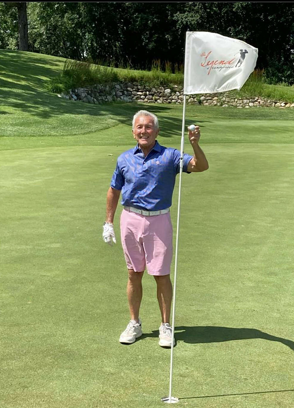 Older man on a golf green holding the flagstick near the hole, wearing a blue shirt and pink shorts under bright sun.