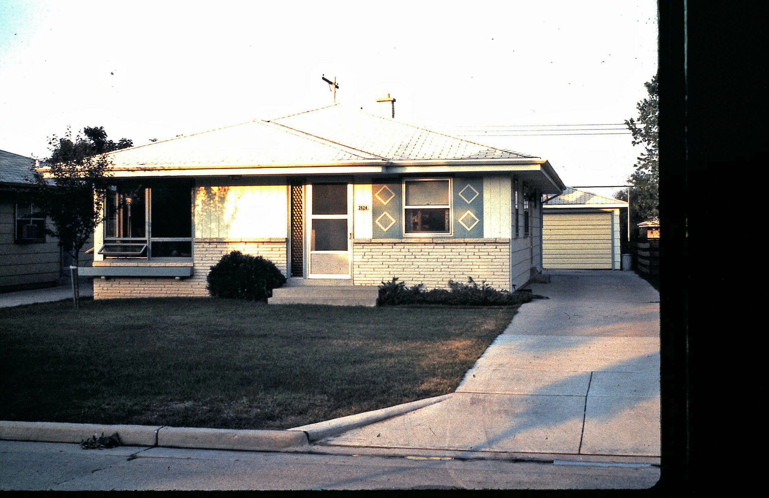 Front view of a single-story suburban house with a brick base, blue facade, and diamond-pattern window accents beside a driveway and lawn.