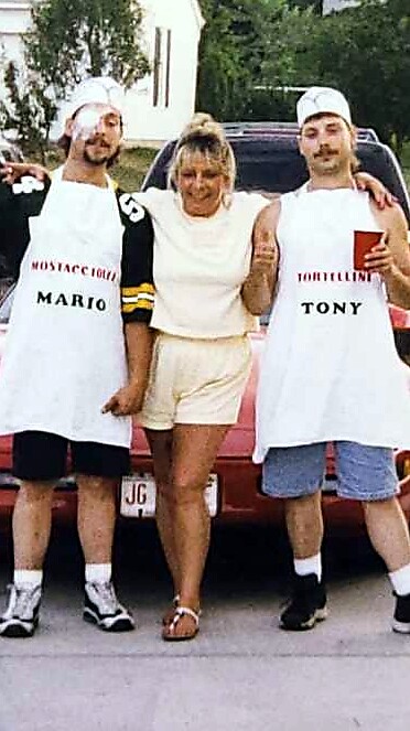 Three people pose together in a parking lot; two men wear aprons labeled Mario and Tony, with a woman between them in a white outfit.