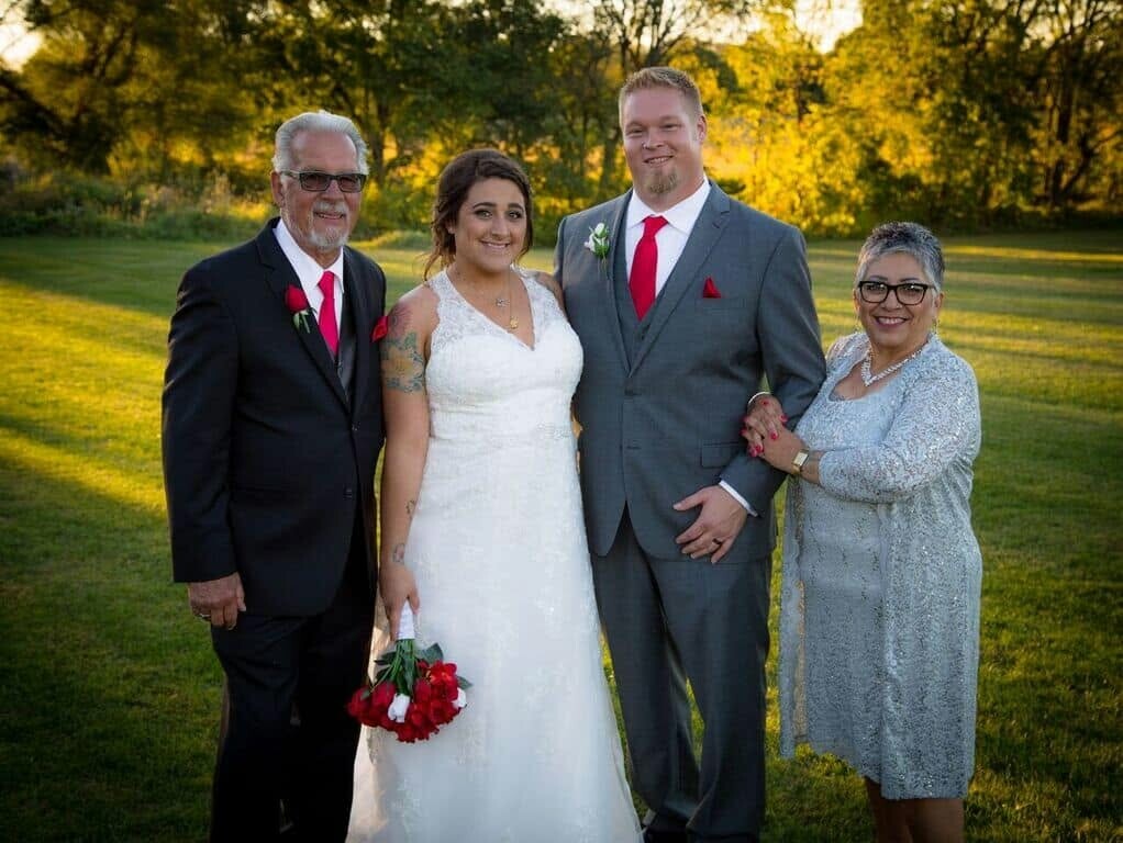 Wedding party outdoors: bride in white gown and bouquet flanked by groom in gray suit and his parents in formal attire at golden hour.
