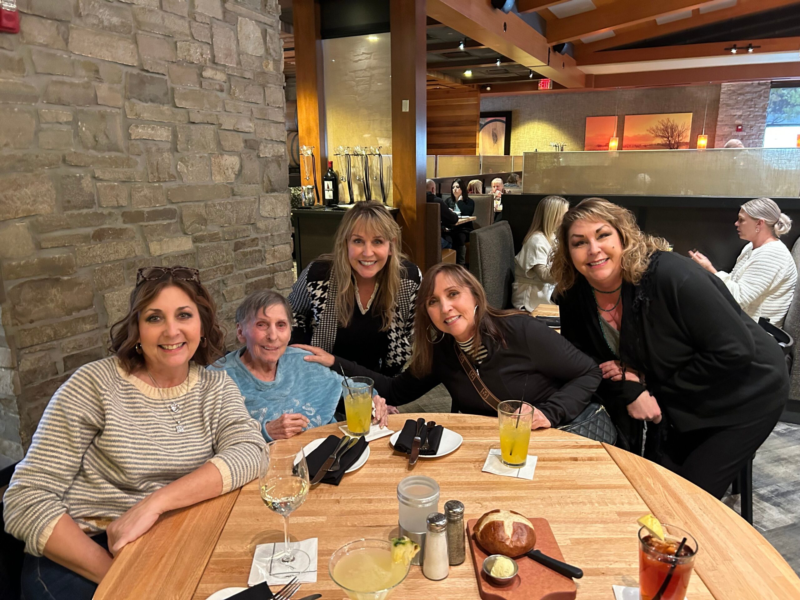 Group of women at a restaurant table smiling for the camera, with drinks, bread, and cutlery on a wooden table and a stone wall in the background.