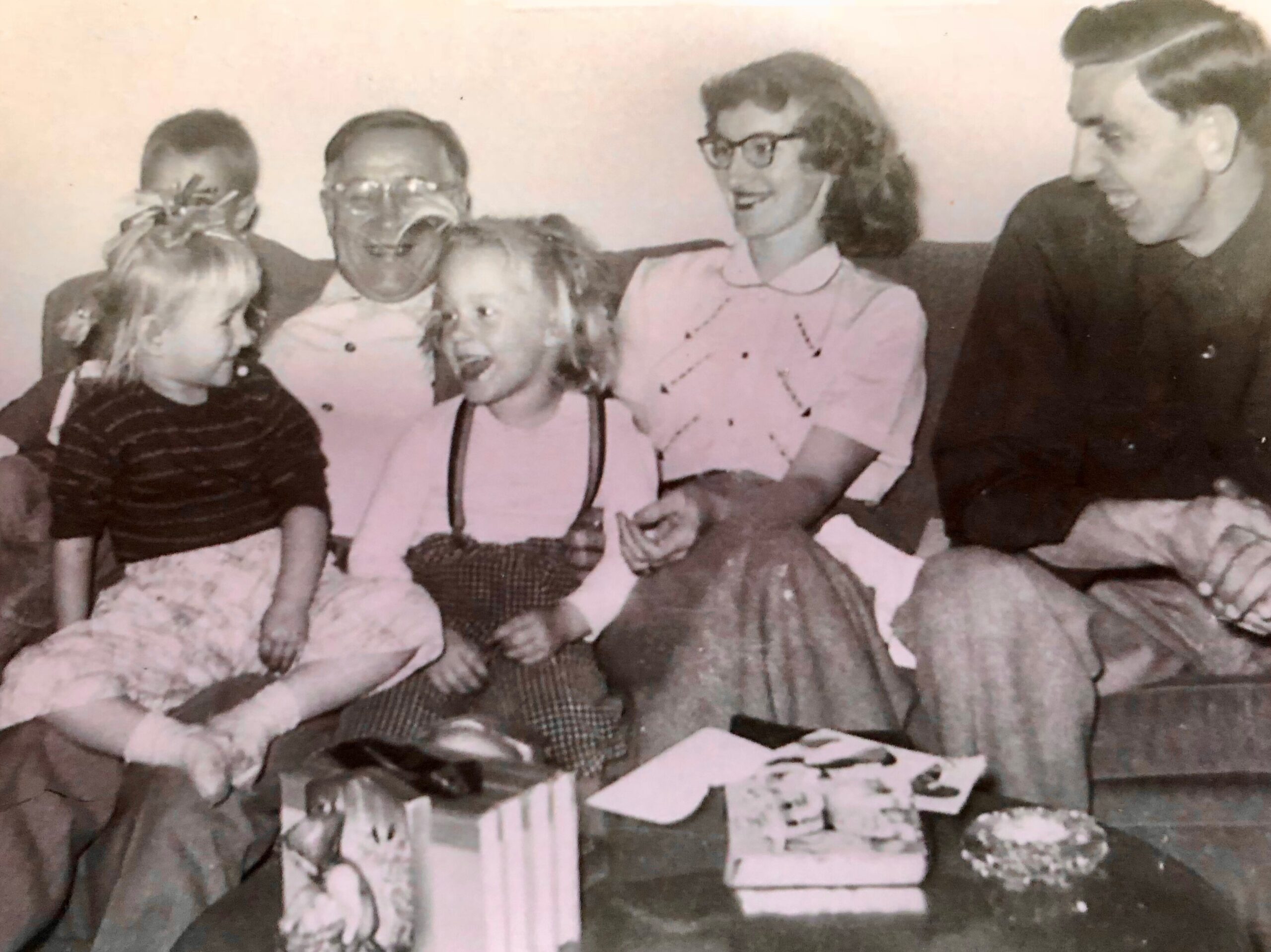 A vintage black-and-white photo of a multiGenerational group of six people sitting on a couch, smiling and gathered around a table with wrapped gifts and a birthday cake visible in front.