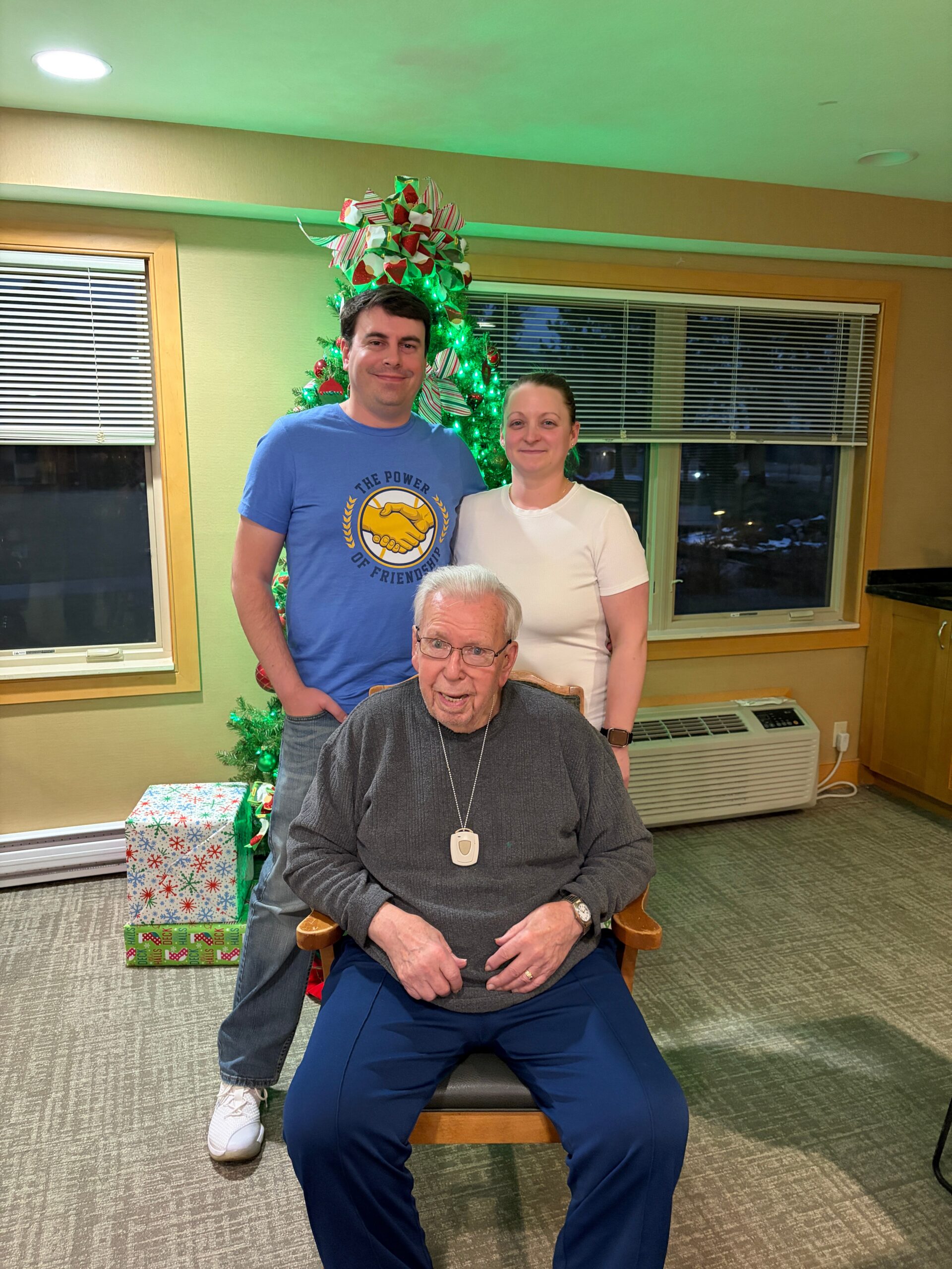 Three people posing indoors by a Christmas tree with wrapped gifts; an elderly man sits in front while a man and a woman stand behind him.