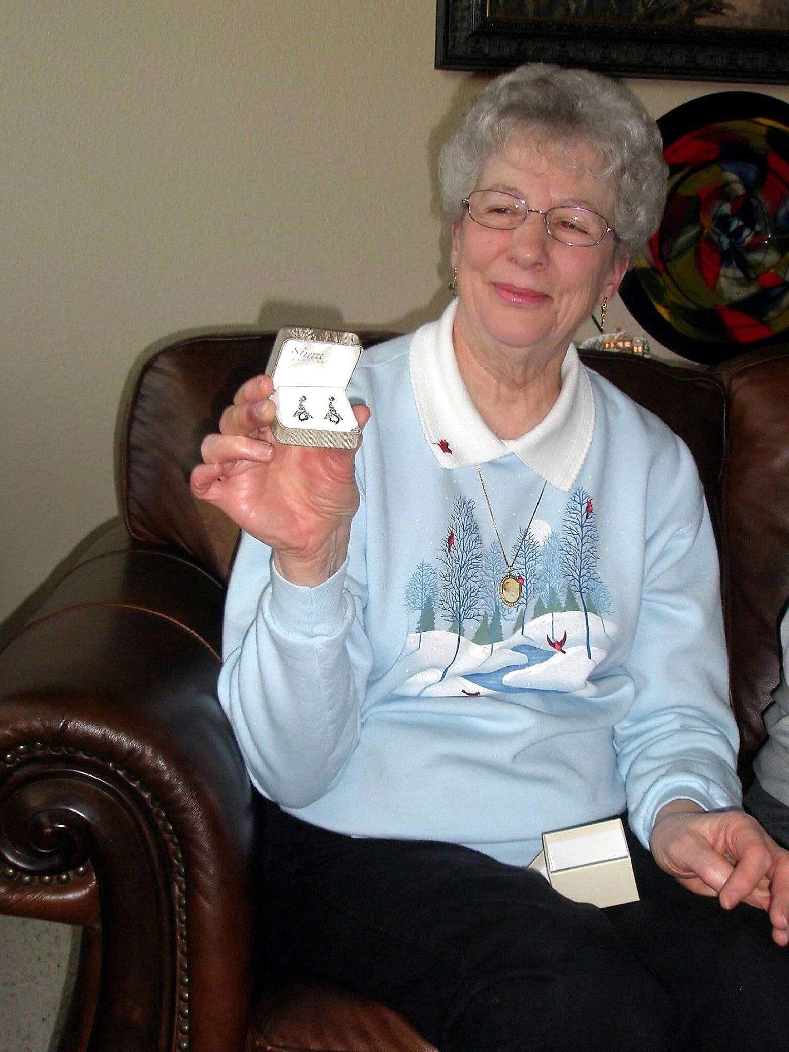 Smiling elderly woman with short gray hair and glasses, presenting an open jewelry box with earrings inside while seated on a leather chair.