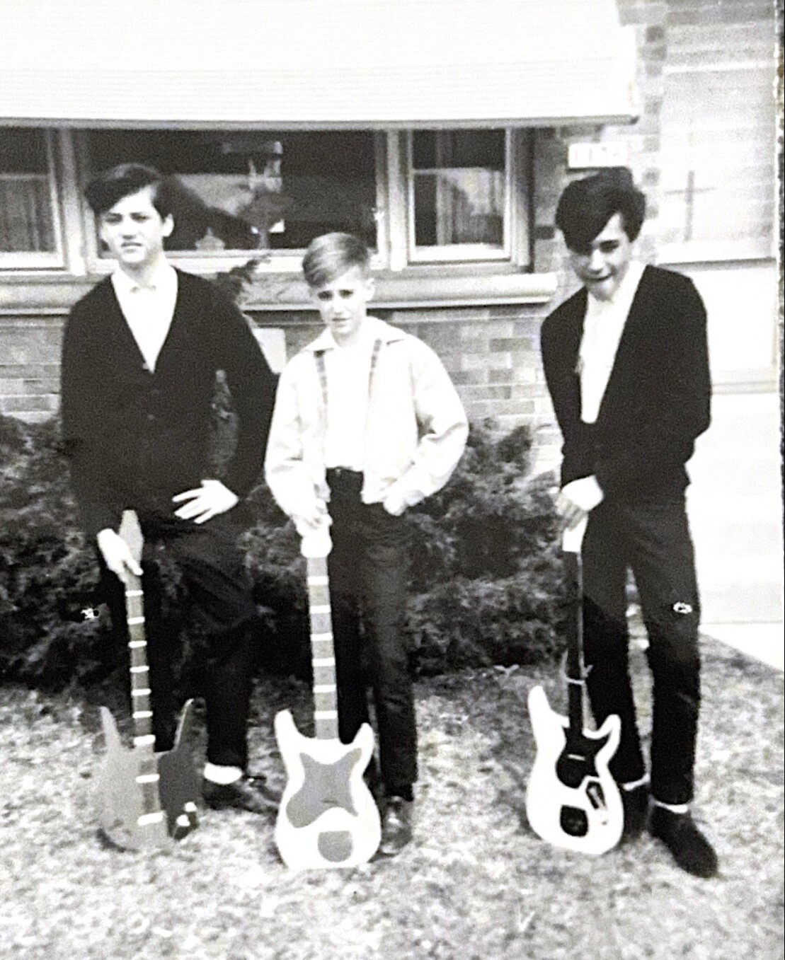 Three young men stand on a lawn in front of a brick house, each with an electric guitar resting on the ground. Left guitarist wears a dark sweater; middle guitarist wears suspenders; right guitarist wears a blazer and smiles slightly.