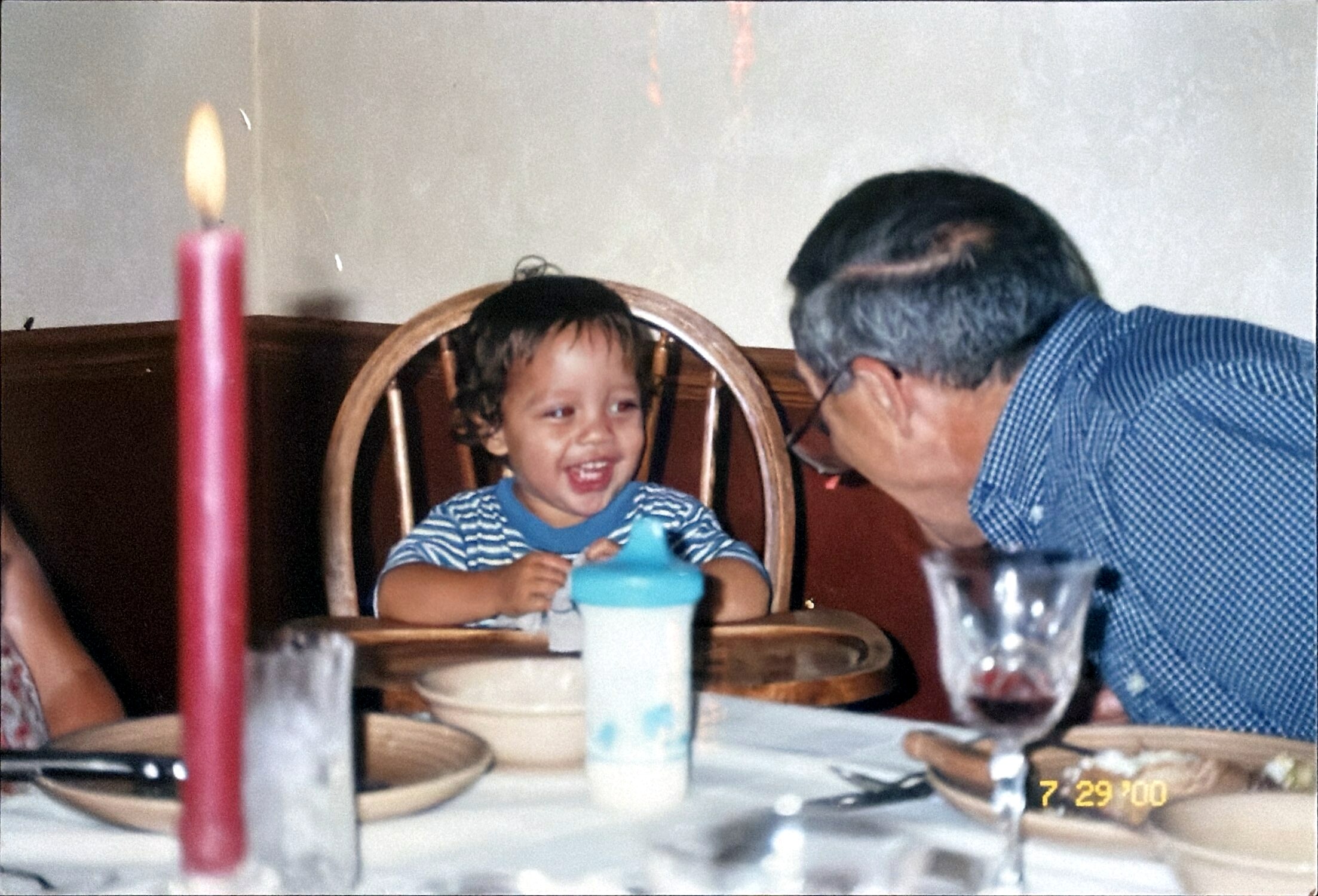 Smiling toddler in a high chair at a birthday table, an adult leaning in nearby; a tall pink candle is in the foreground on the left.
