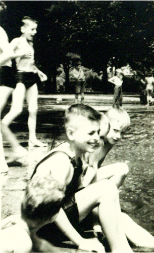 Children in swimsuits sit along the edge of a park pool or pond, smiling, with others standing in the background.