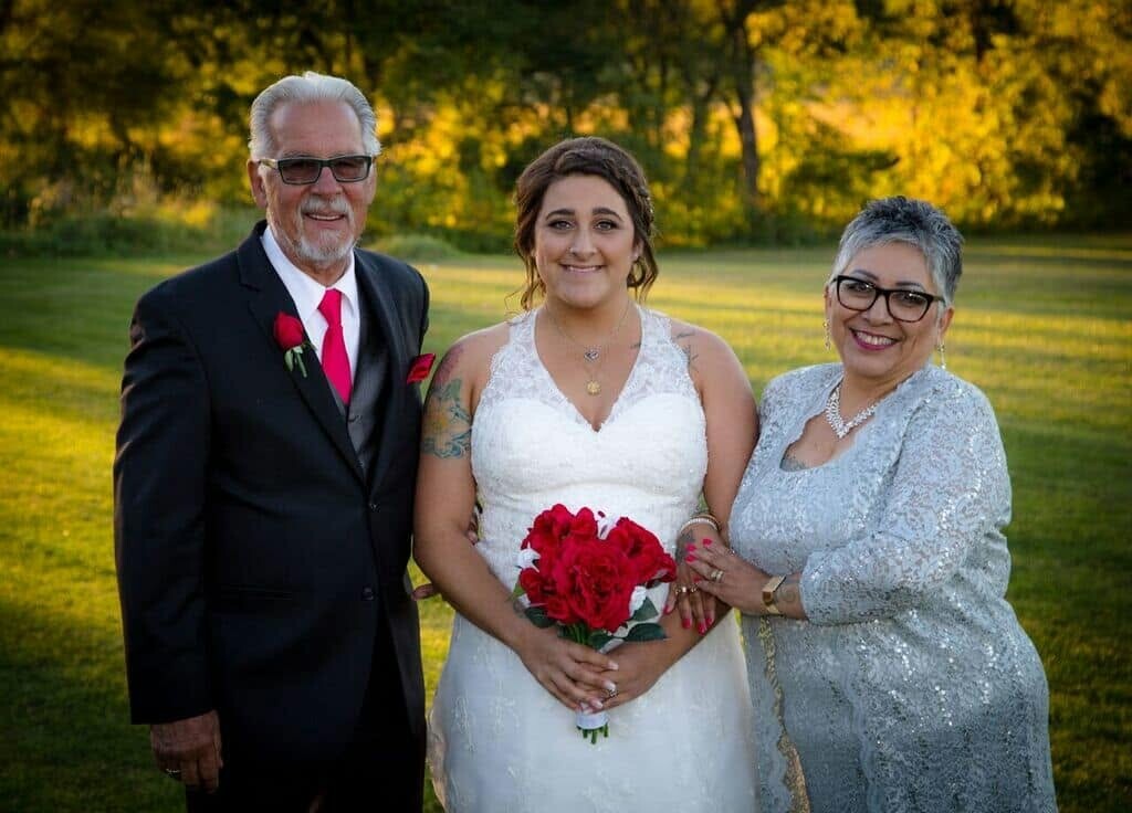 Bride in a white lace gown holding a red bouquet, flanked by an older man in a black suit and a silver-haired woman in a sparkling silver dress on a sunny lawn.