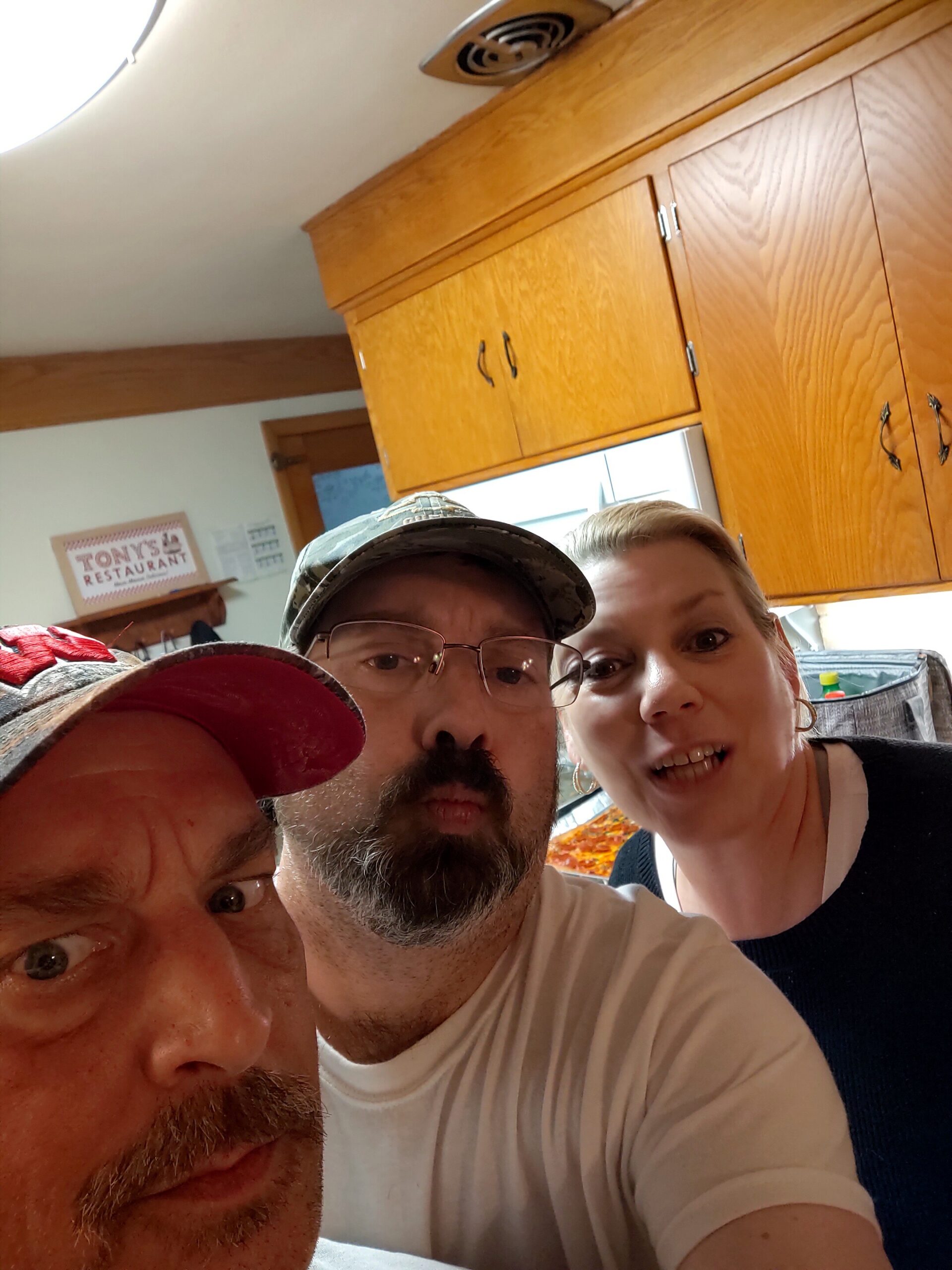 Three people are taking a close-up selfie in a kitchen with wooden cabinets in the background, one smiling person at right.