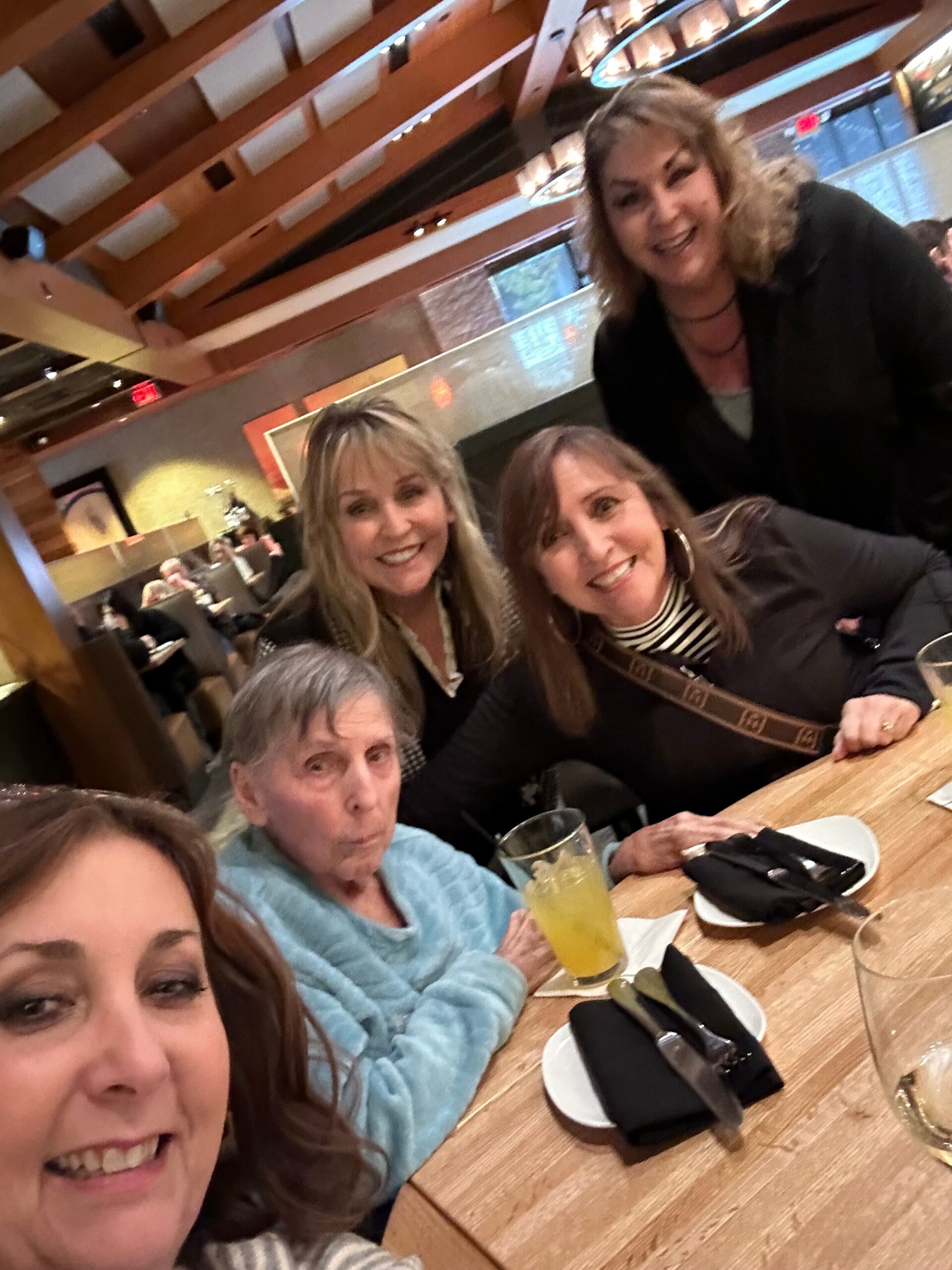 Group of friends smiling around a wooden restaurant table, taking a selfie together in a casual dining setting with drinks and plates nearby.