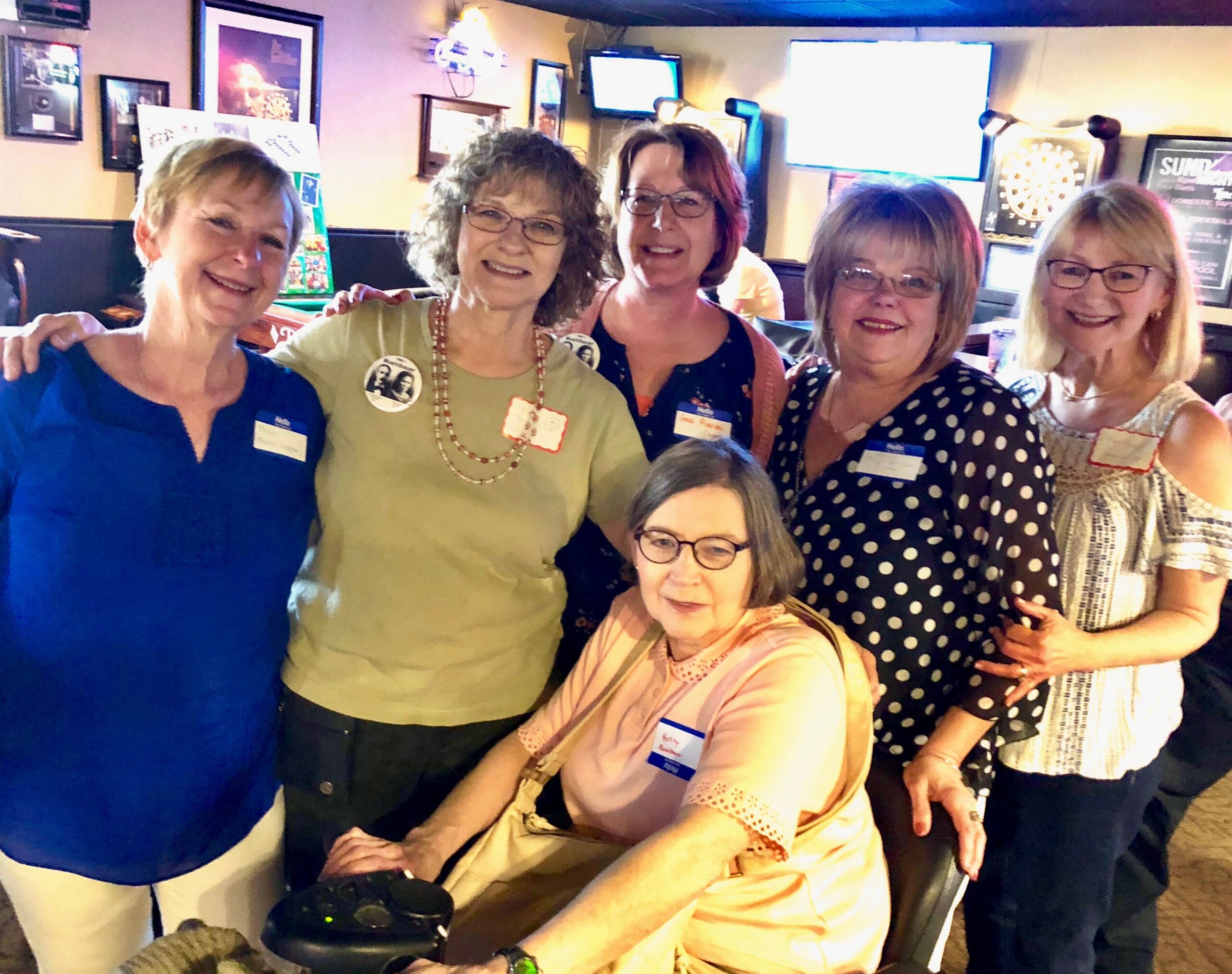 Five smiling women posing for a group photo in a bar, wearing name badges and casual outfits, darts board and televisions in the background.