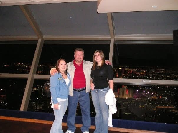 Three adults pose together on a nighttime observation deck with city lights in the background; man in red polo and light jacket between two women.