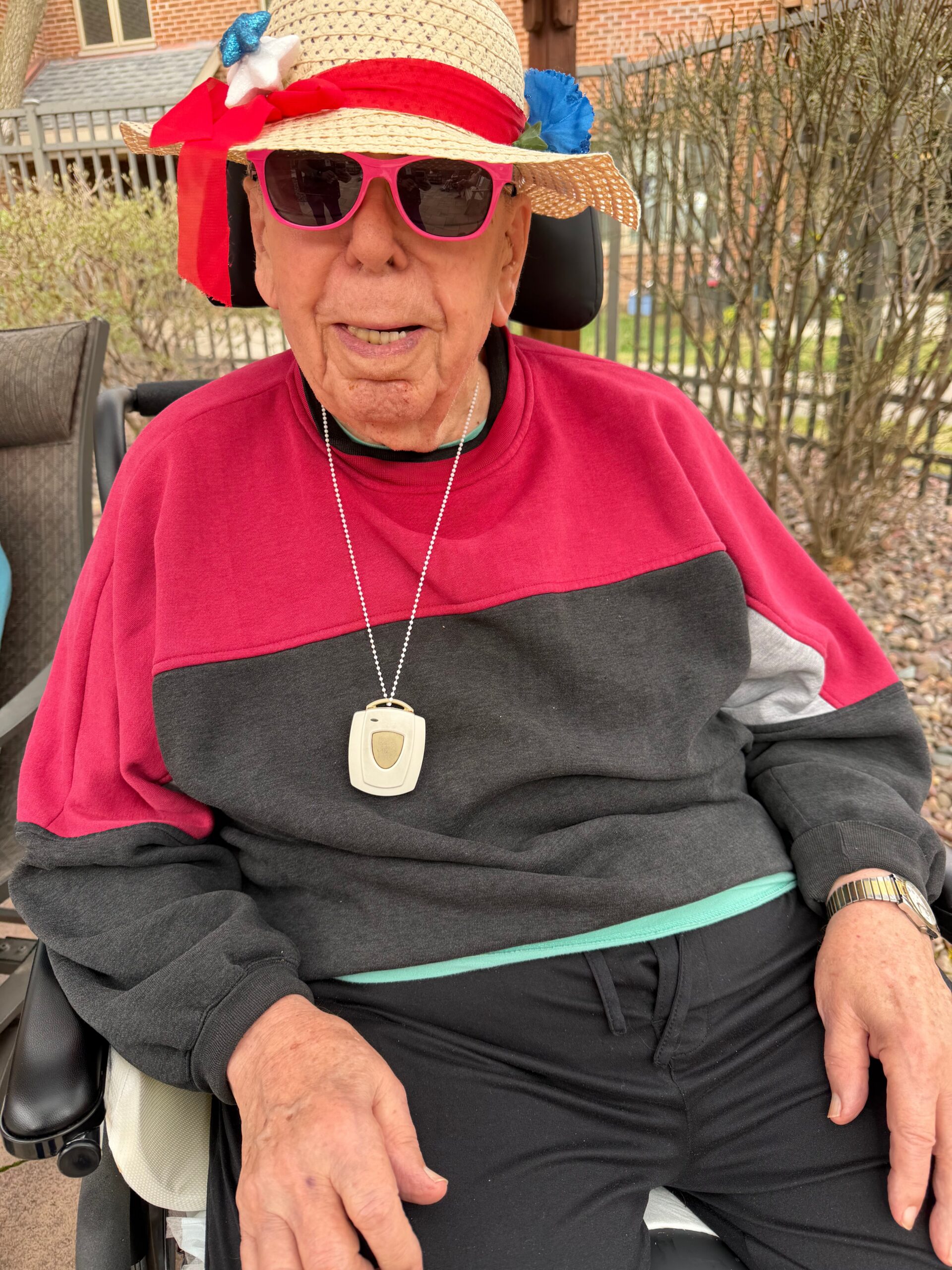 Elderly person in a wheelchair wearing a straw hat with a red ribbon, pink sunglasses, and a red-and-black top outdoors.