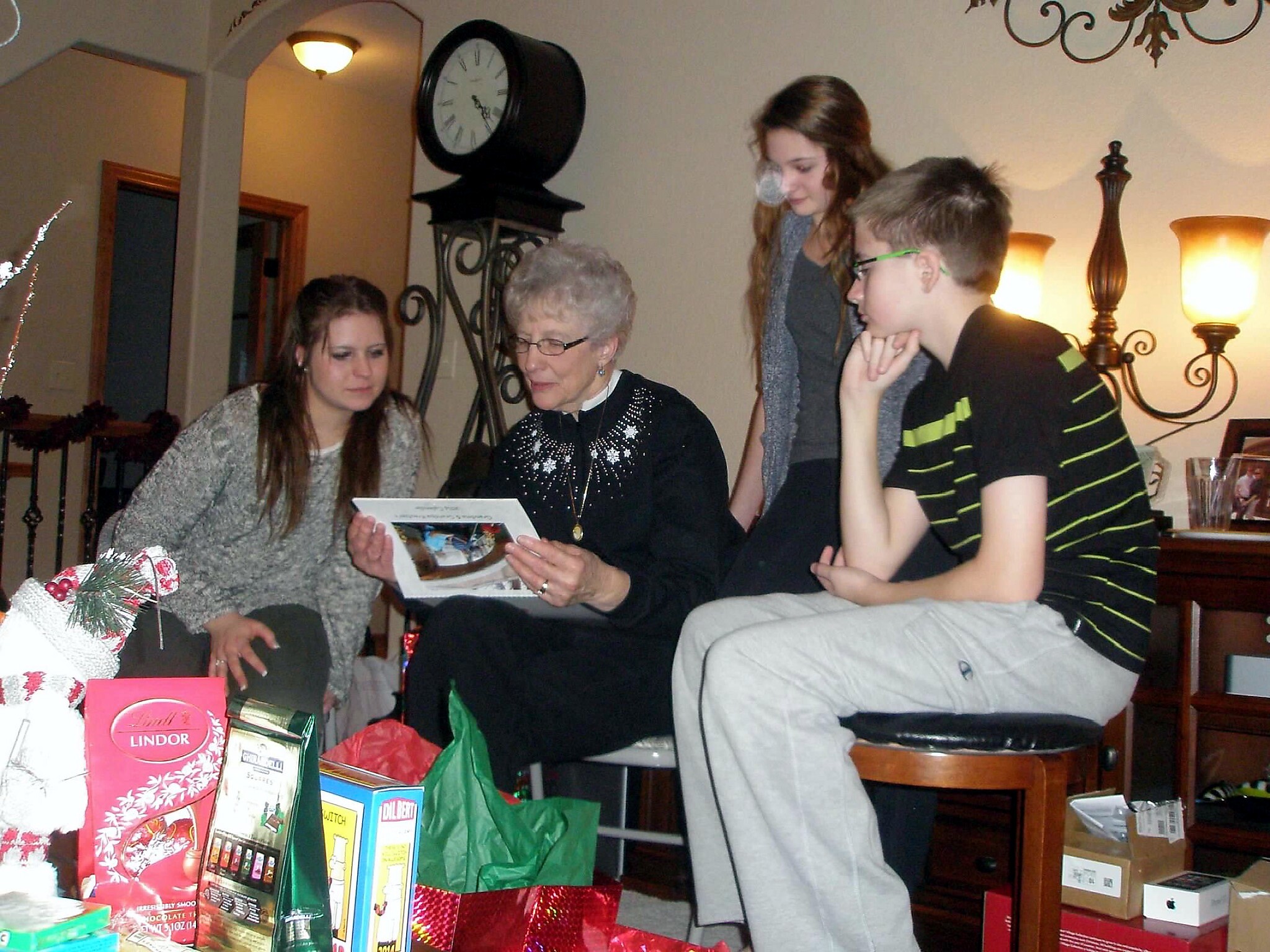 Older woman reads a card with younger relatives nearby during a holiday gift exchange, with wrapped presents around them in a living room.